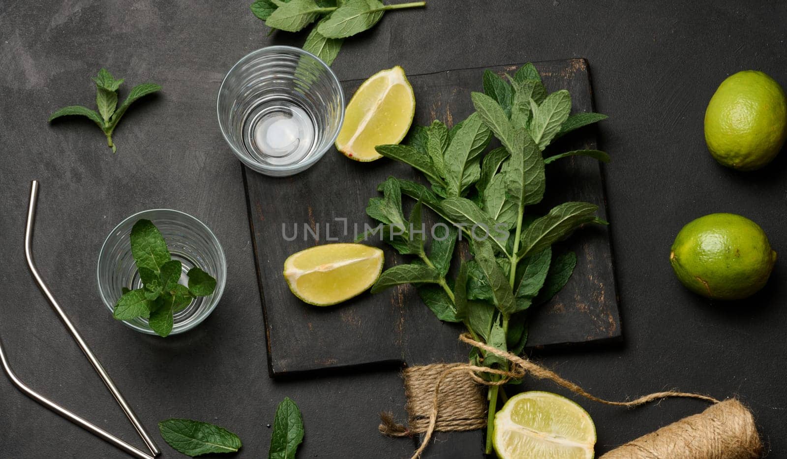 Bunch of fresh green mint on wooden board and lime wedges, glasses and metal straws, top view. Cocktail ingredient