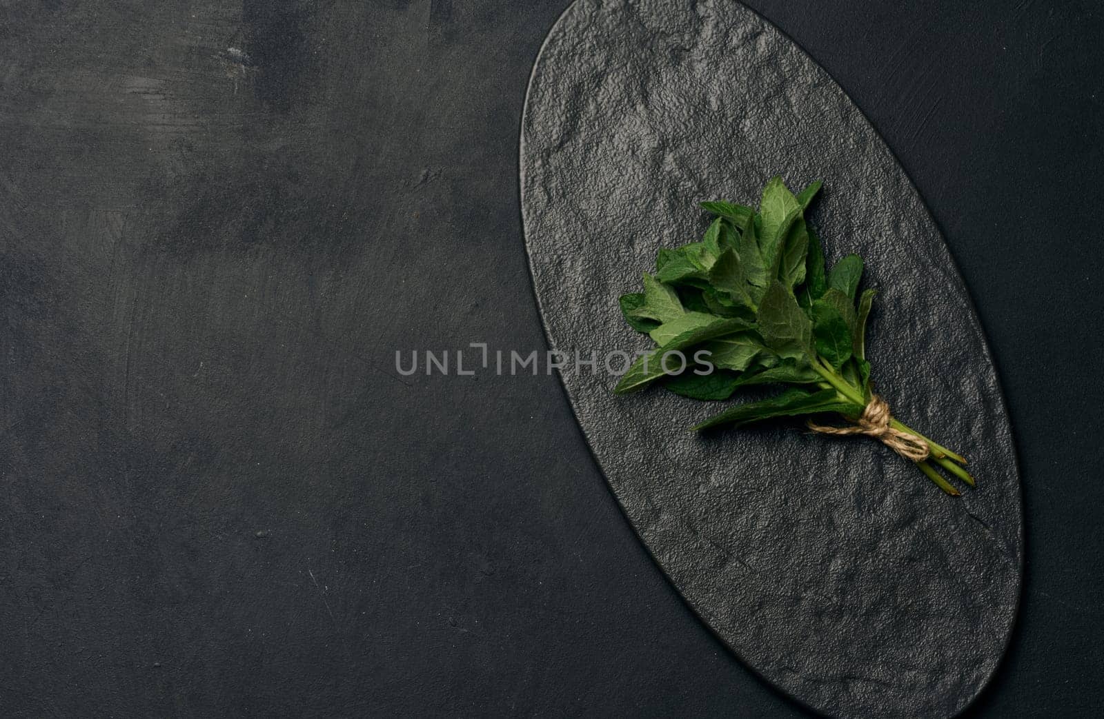A small bunch of fresh green mint sprigs tied with brown twine rests on an empty, textured black oval plate. Copy space