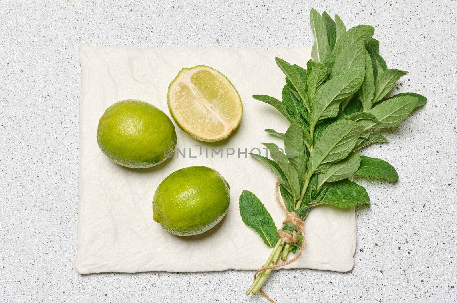 Whole limes and a halved lime are arranged next to a small bunch of fresh mint leaves tied with twine, top view
