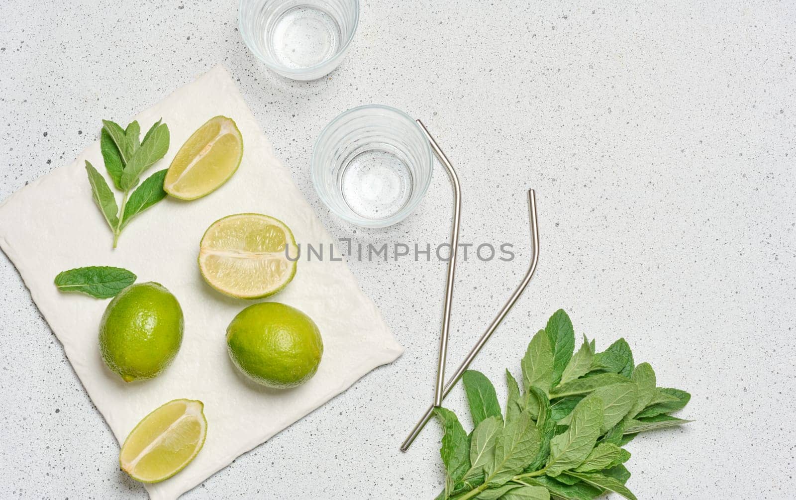 A top down view of ingredients for a refreshing drink, including whole and sliced limes, fresh mint sprigs, two empty glasses, and two metal straws