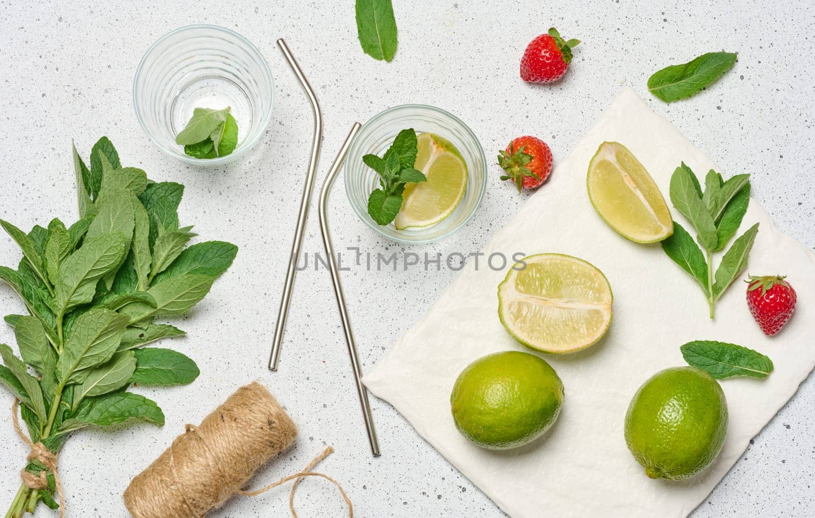 A top down view of ingredients for a refreshing drink, including whole and sliced limes, fresh mint sprigs, two empty glasses, and two metal straws