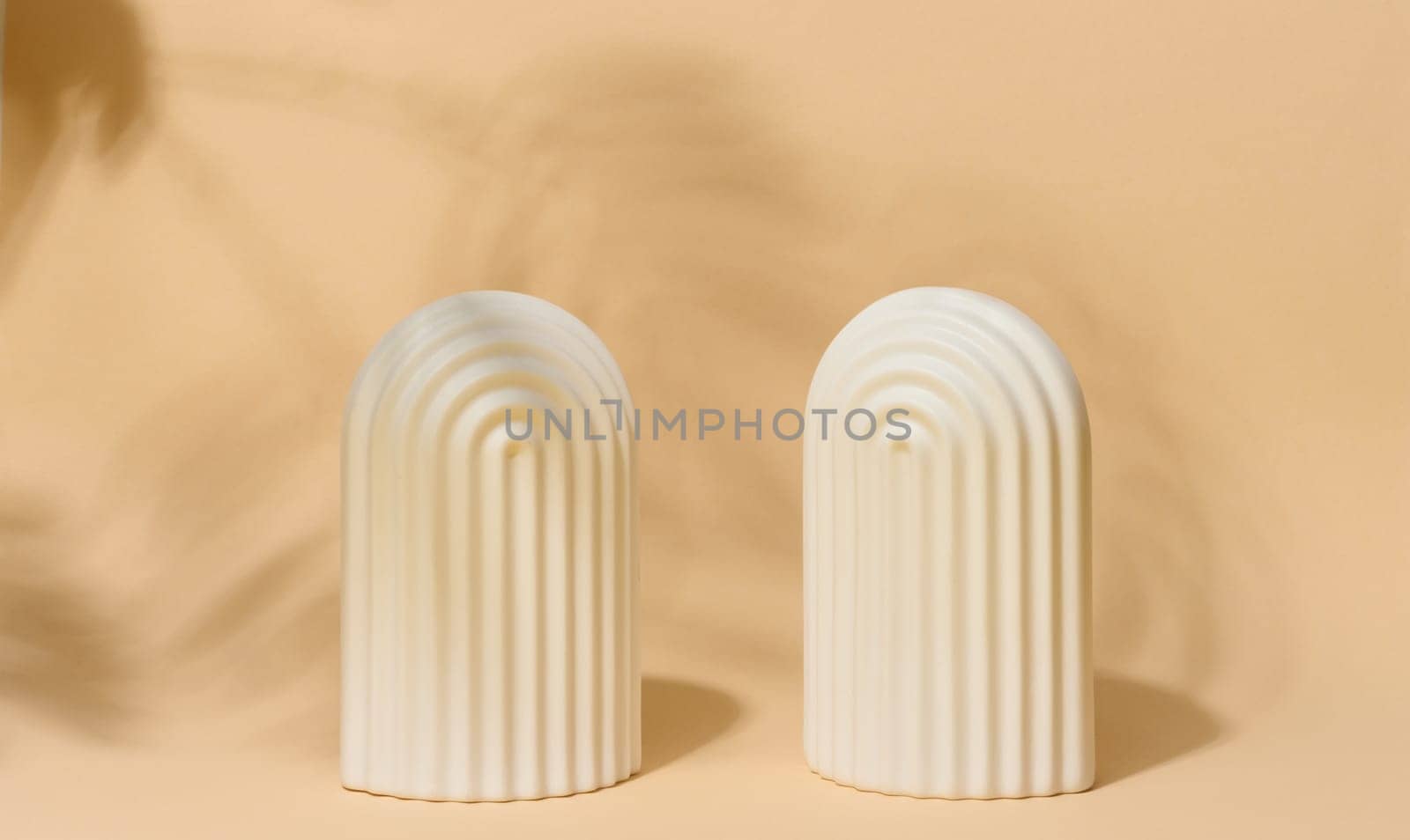 A small, white ceramic decorative arch with vertical ribbed texture stands on a beige surface with blurred leaf shadows