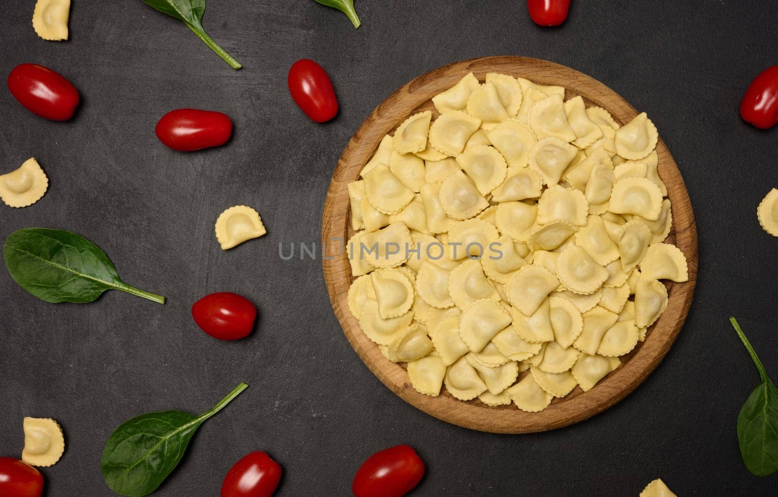 Uncooked tortellini pasta with fresh cherry tomatoes and spinach leaves on a dark, textured surface. Flat lay 