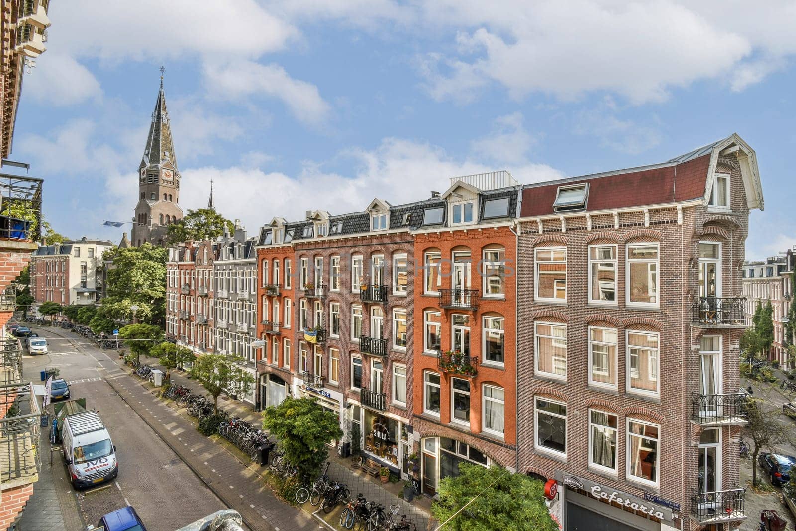 A picturesque view of an Amsterdam street featuring traditional brick buildings, trees lining the road, and a stunning church spire in the background.