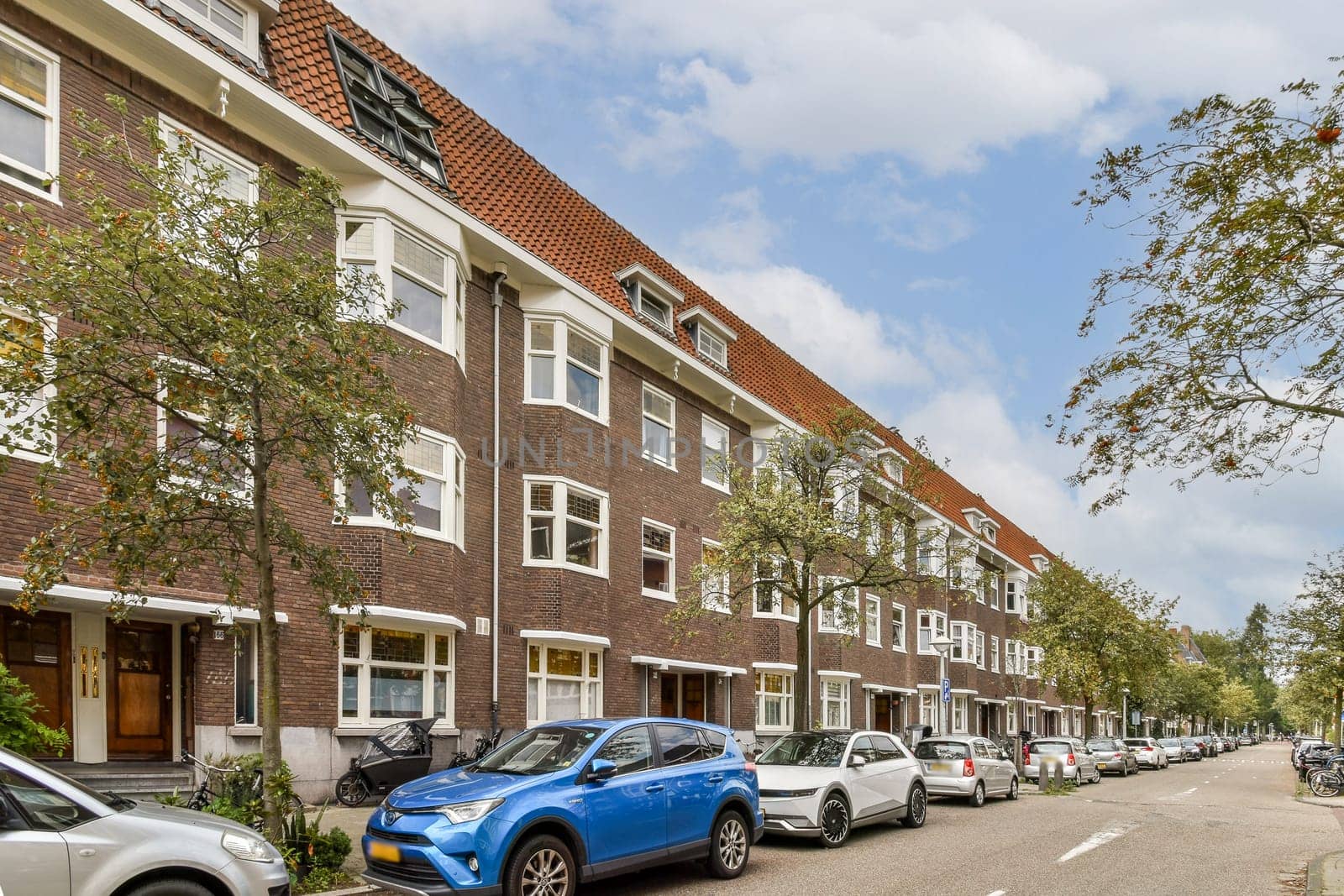A picturesque view of a residential street showcasing diverse architectural styles and well-maintained greenery. Various parked cars line the street, enhancing the urban charm.