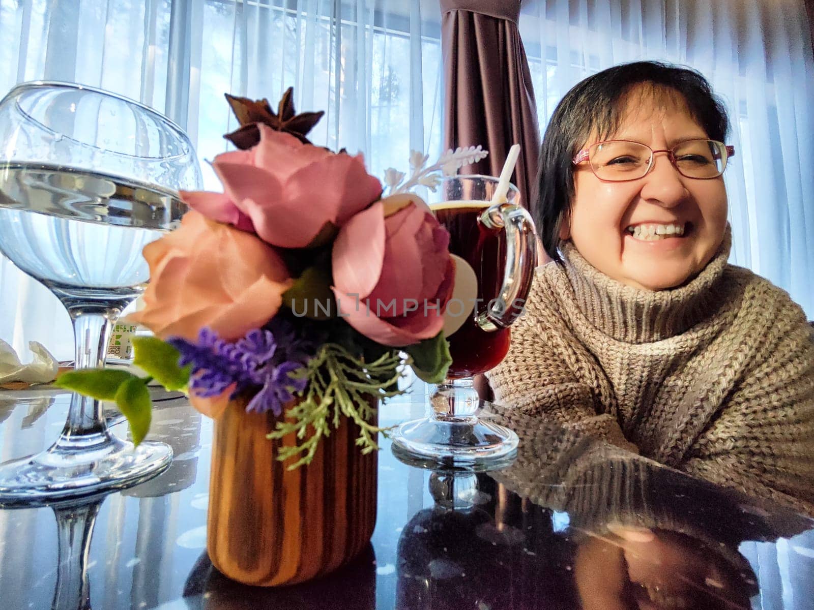 A woman smiles joyfully while sitting at a cozy cafe table. She enjoys a beverage, surrounded by a small floral arrangement, with soft light filtering through nearby curtains.