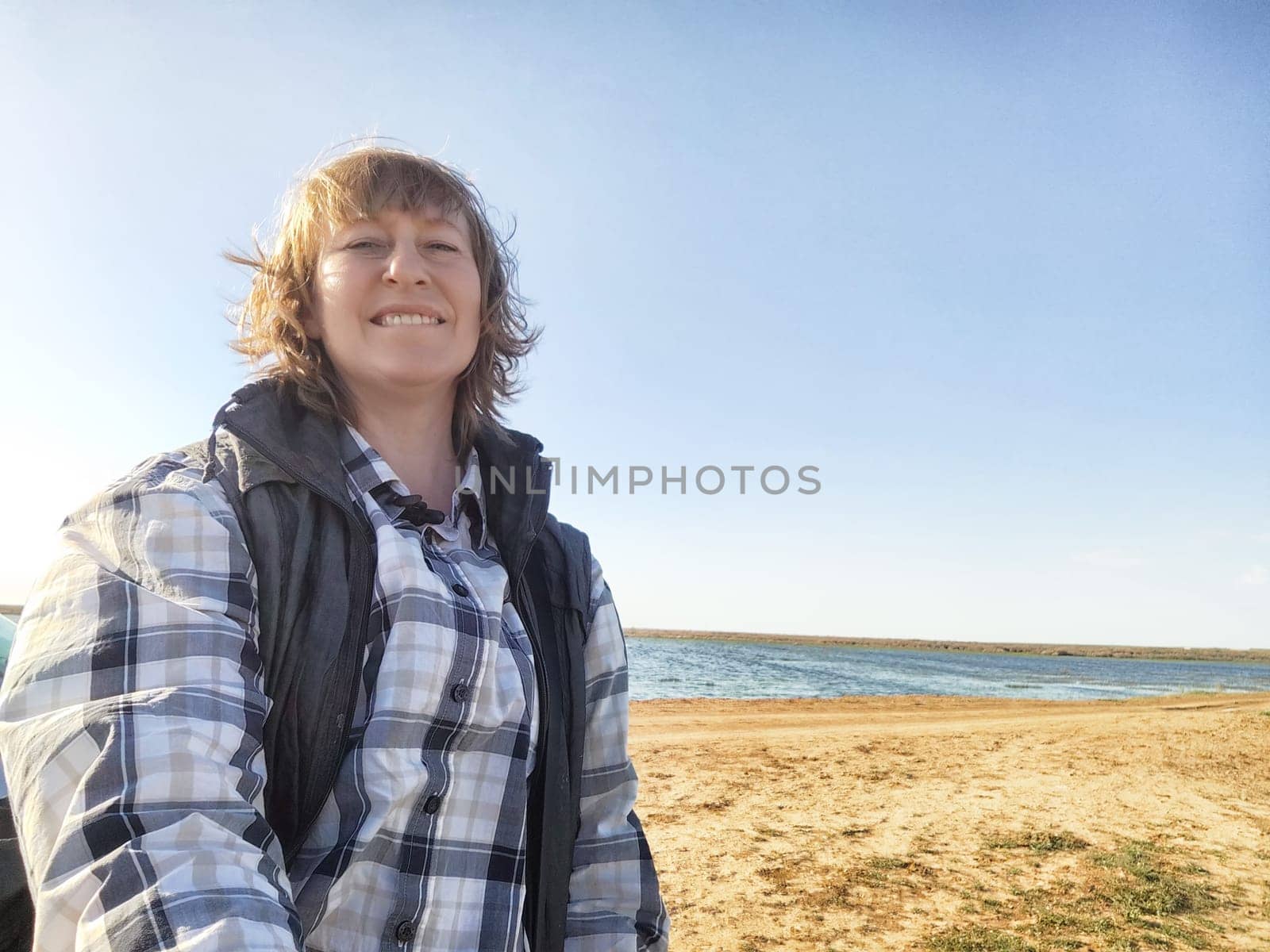 Blonde adult enjoys a leisurely trip at a scenic beach, smiling for a selfie with the water in the background under a clear sky.