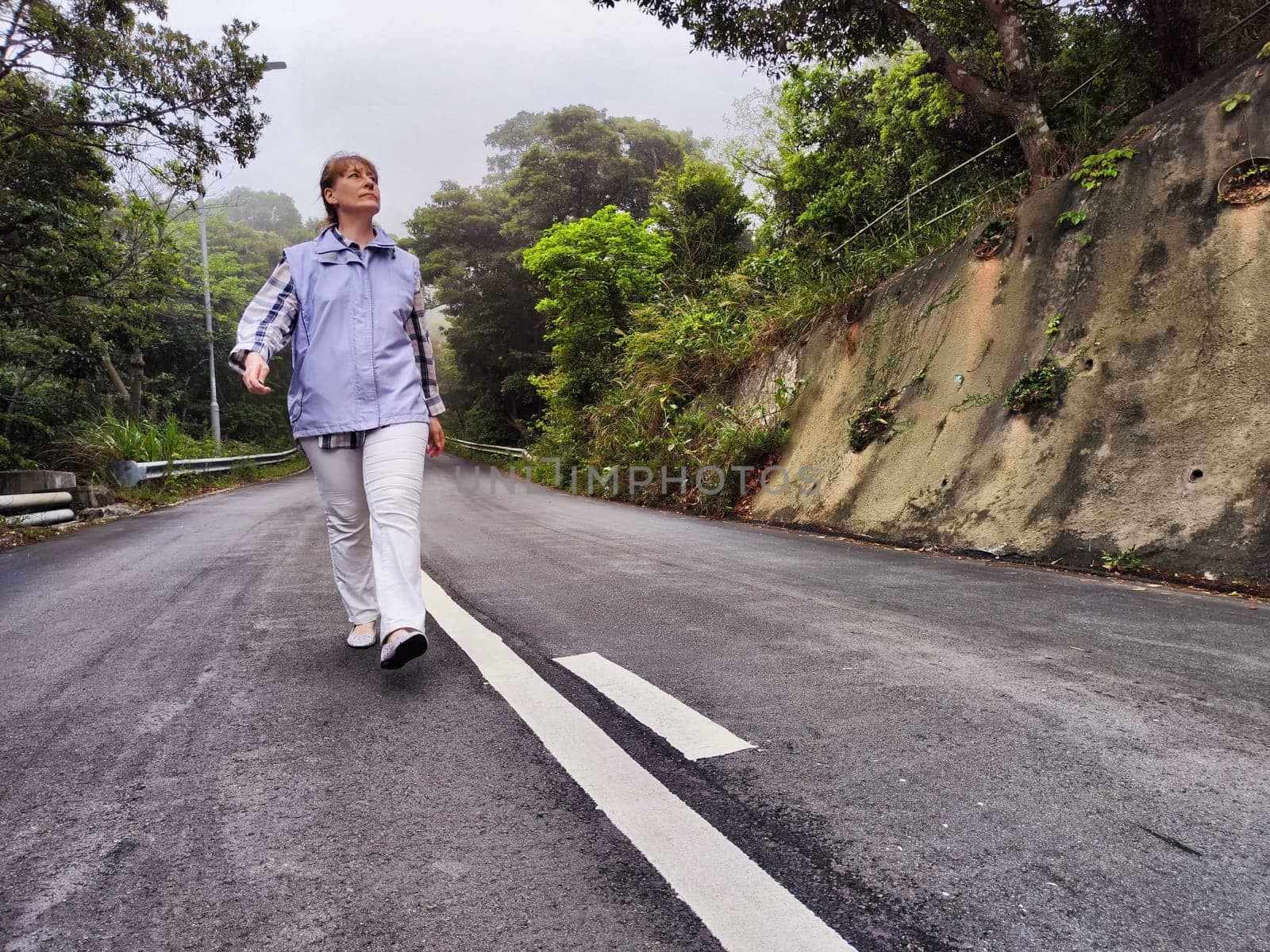 A mature woman strolls confidently down a tranquil road, showcasing her relaxed lifestyle amidst lush trees on an overcast day.