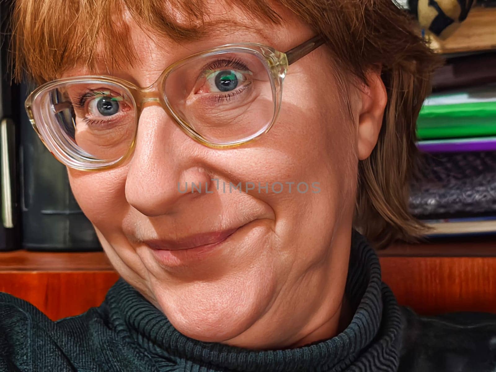 A mature woman smiles brightly while taking a selfie in a warm, inviting indoor space filled with books and personal items reflecting her lifestyle.