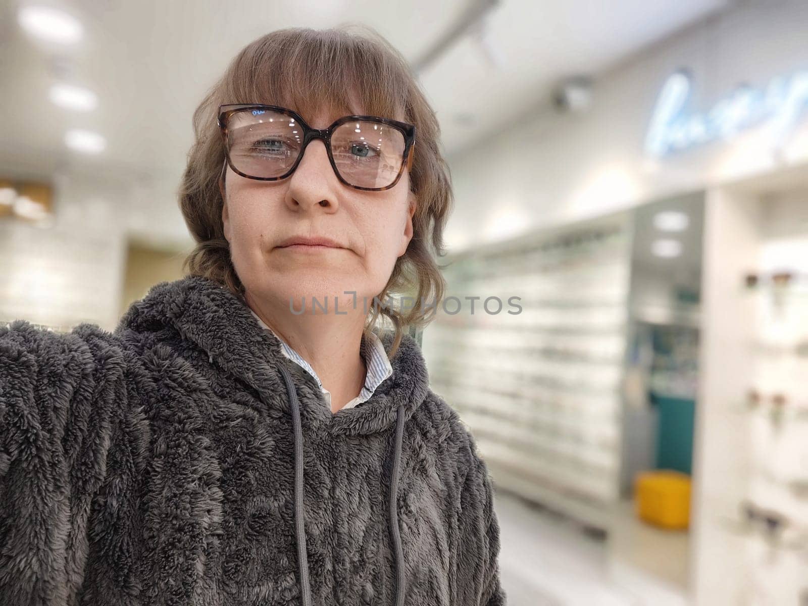 A middle-aged woman with blonde hair evaluates her appearance while trying on glasses in an optics store filled with various options.