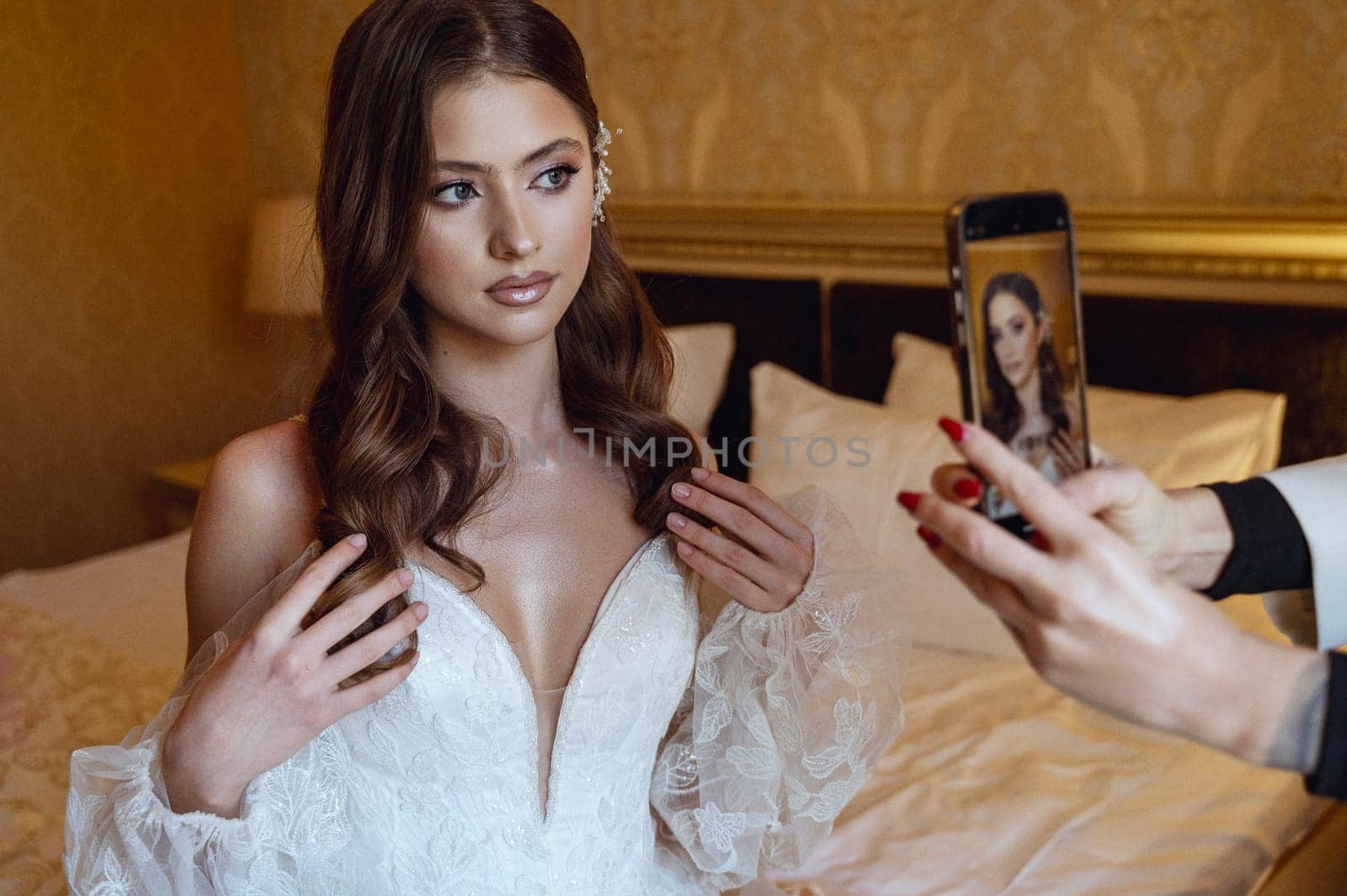 Bridesmaid taking photos of beautiful bride on her phone. Bride with makeup and hairdo in lace dress posing in front of phone camera. Wedding day morning.