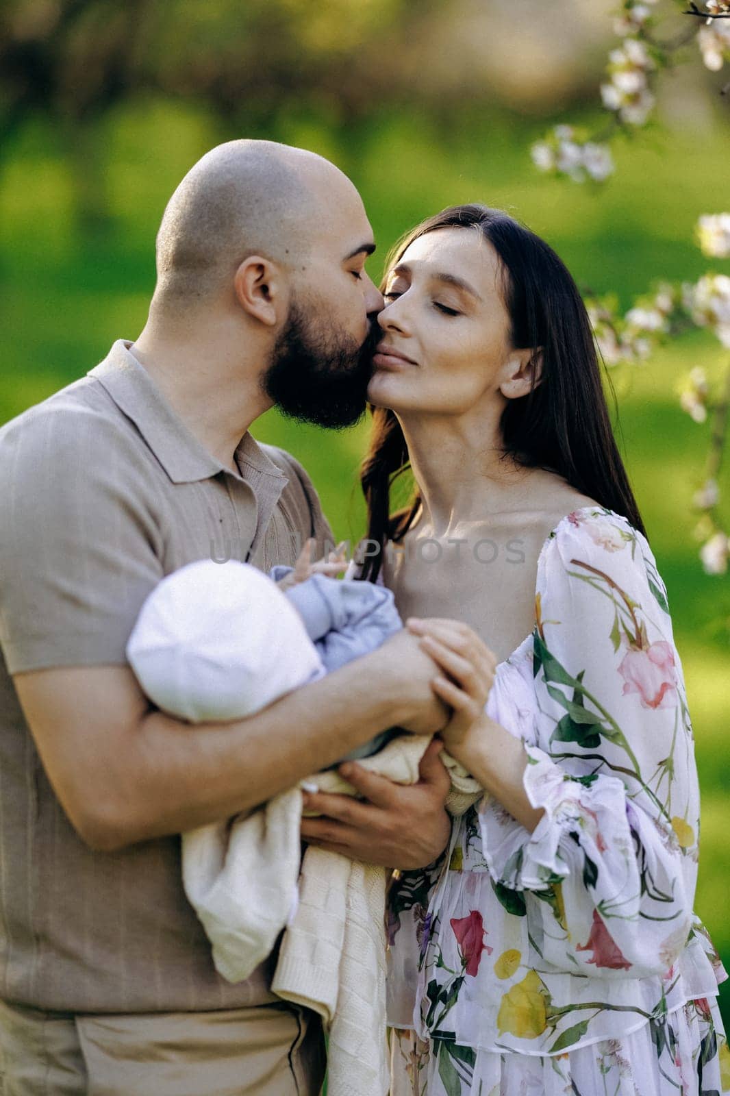 A man and a woman are gently holding a baby together in a beautiful park filled with blooming flowers and lush greenery around them
