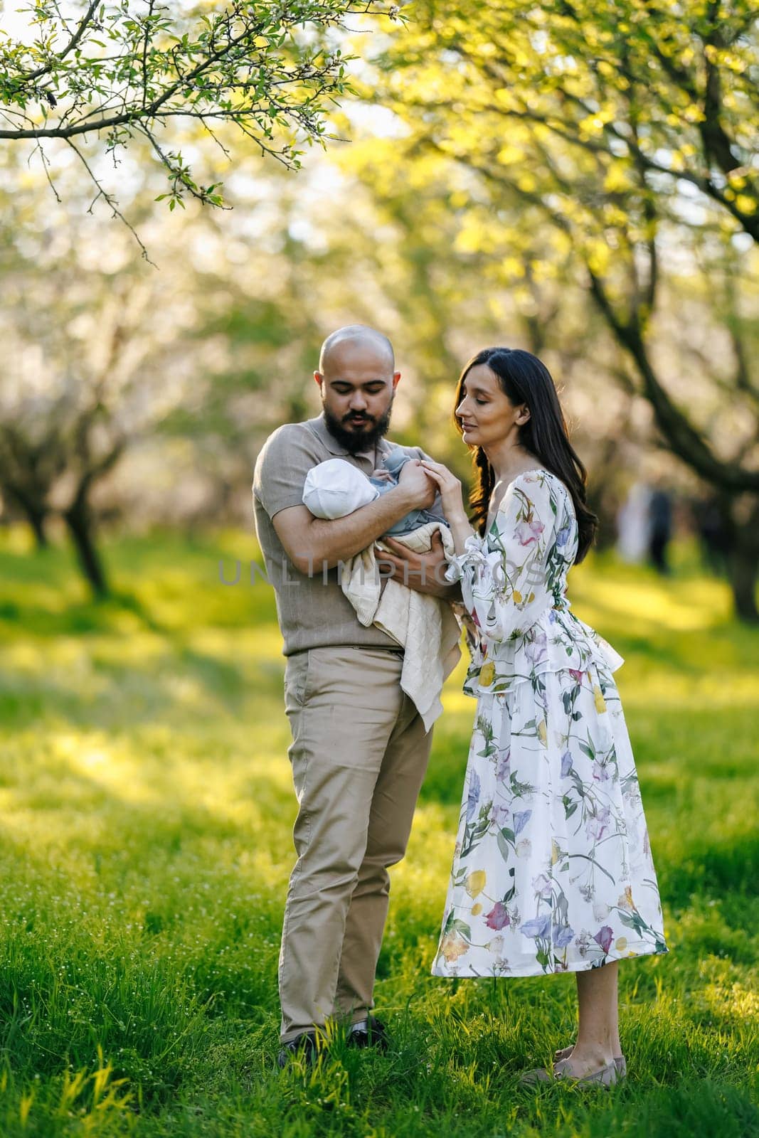 In a lovely park filled with greenery, a man and a woman are gently holding a cute baby together, creating a memorable moment