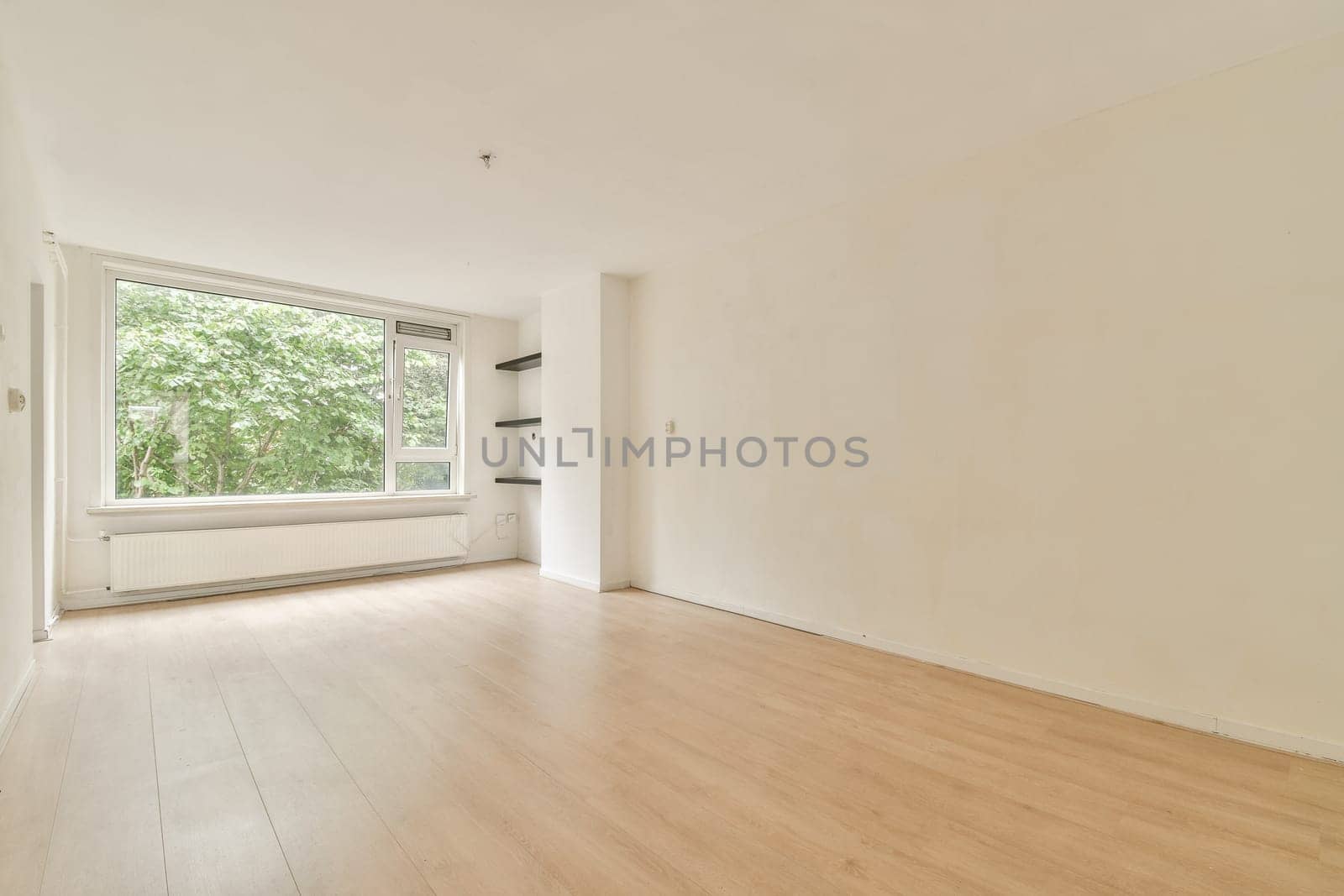 An empty, minimalist interior room featuring large windows allowing natural light to fill the space. The wooden floor complements the bright atmosphere.