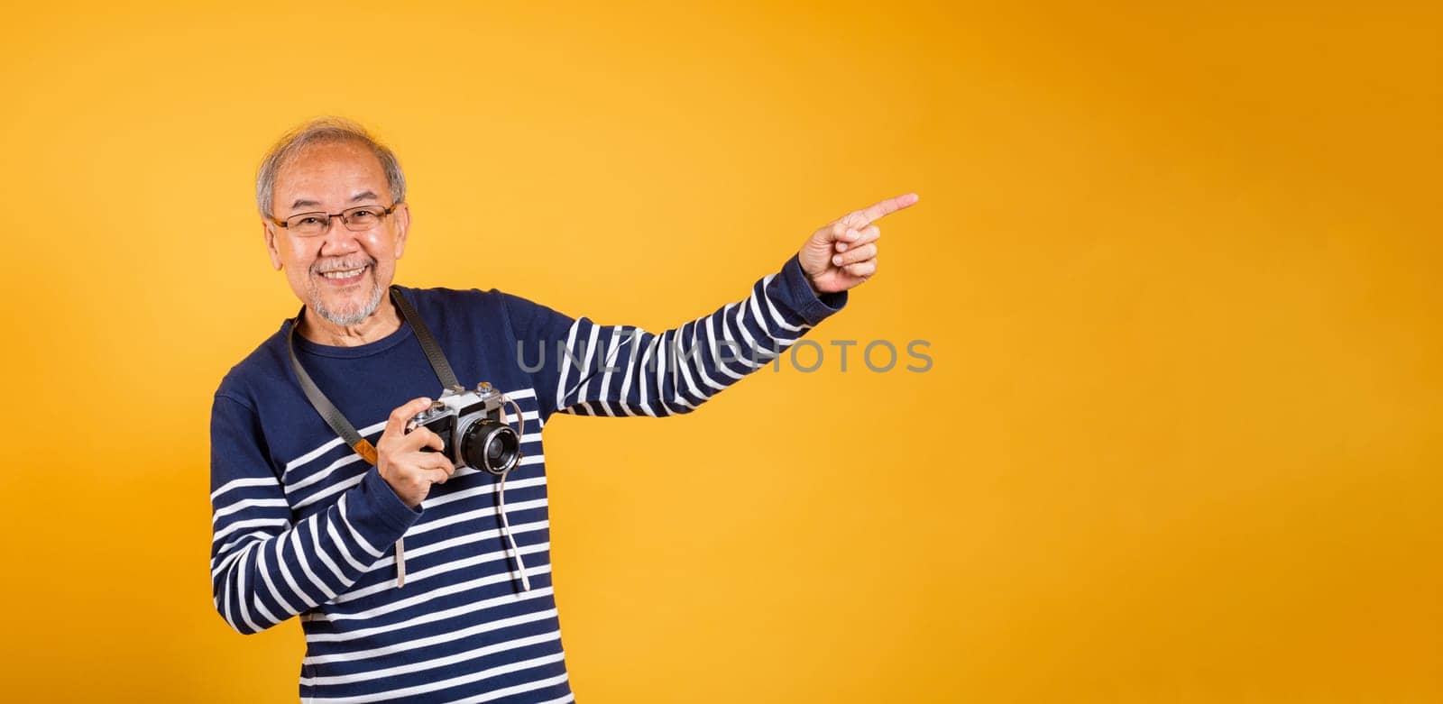 Portrait Asian old man wearing glasses hold vintage SLR camera and pointing something side away studio shot isolated yellow background, smiling happy Photographer elderly man gray haired