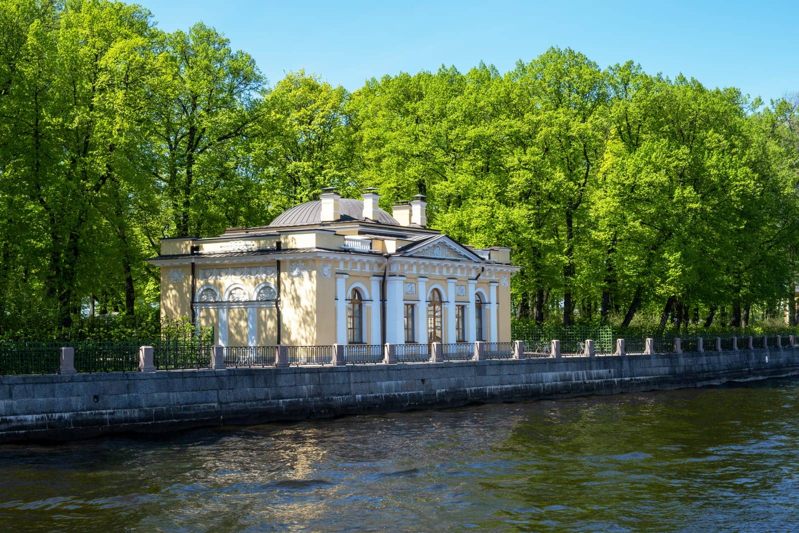 Saint-Petersburg, Russia - 05.21.24. Historic riverside building with classical architecture set against vibrant green trees of Summer garden. Showcasing serene water and scenic natural surroundings