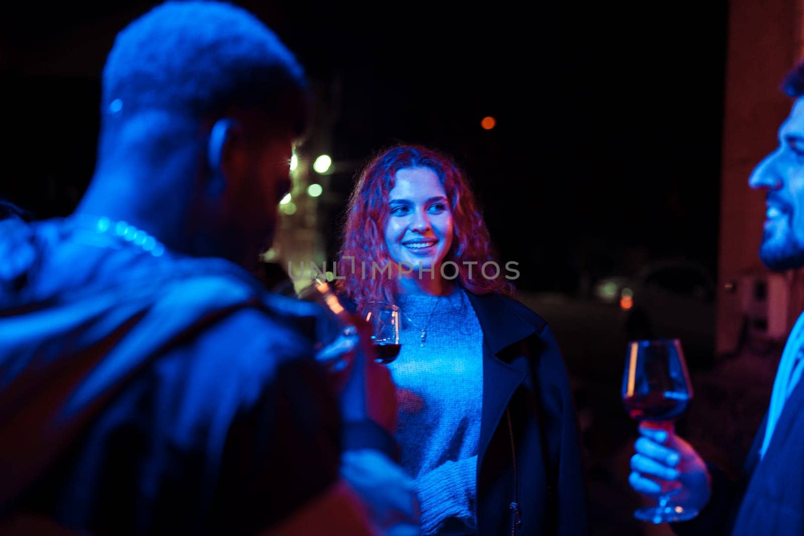 Three friends converse and share laughter while holding glasses of wine during an evening social event. The vibrant blue and red lights create a festive atmosphere.