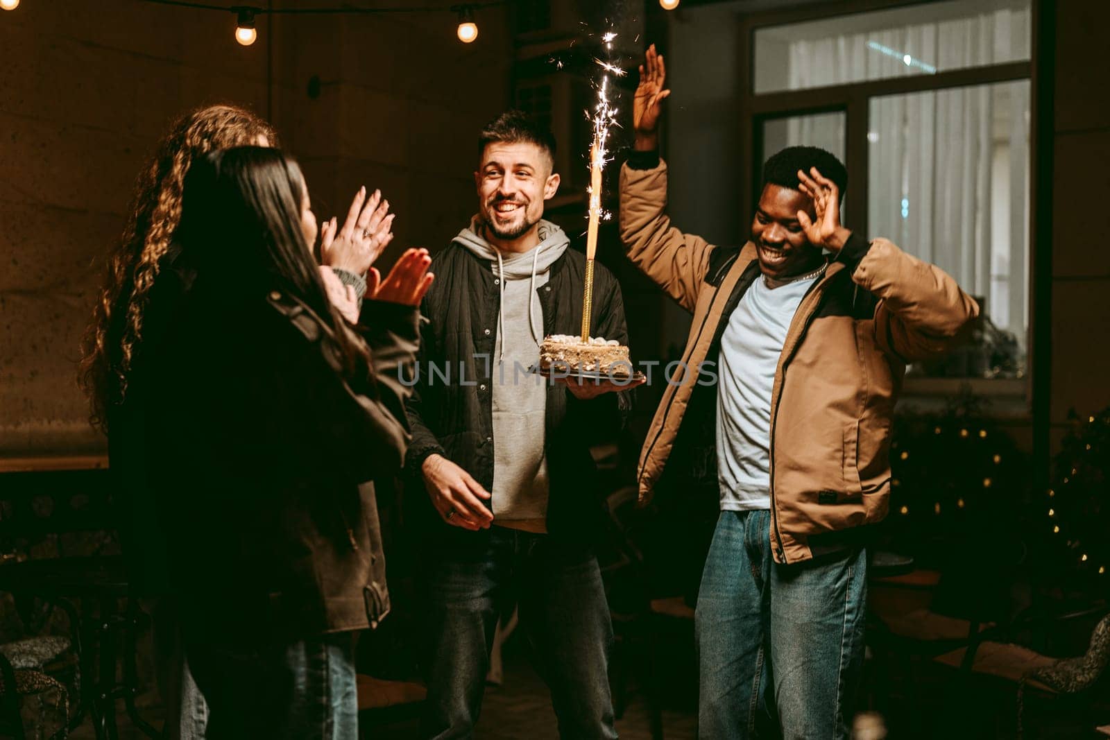Friends gather in a cozy night setting, celebrating a special occasion as one person holds a birthday cake adorned with sparklers. Laughter and cheers fill the air.