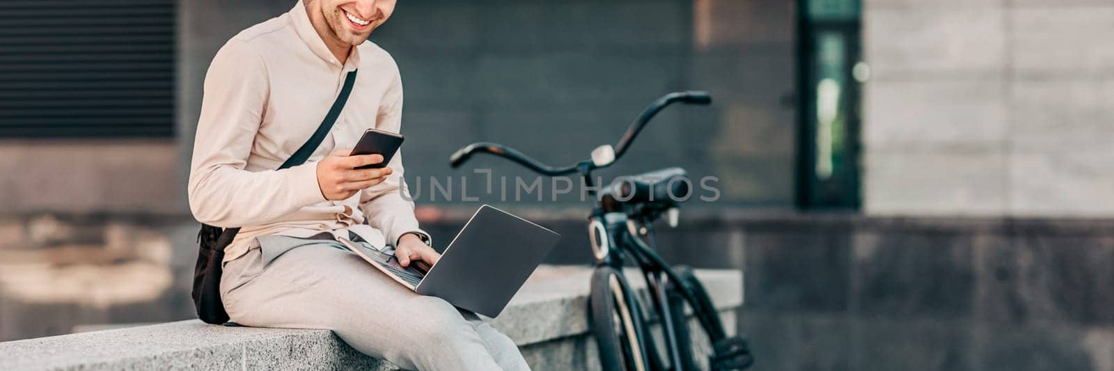A young man smiles while checking his smartphone, sitting on a concrete wall with his laptop beside him. His bicycle rests close by, capturing a casual urban lifestyle moment, copy space