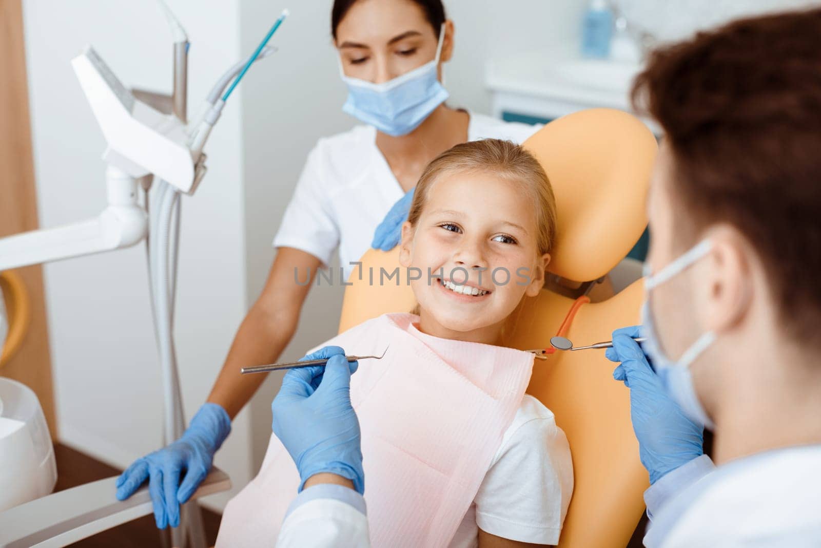 Modern dental examination and oral cavity treatment in children. Cheerful little kid in medical chair looks at millennial male doctor in white coat, protective mask and rubber gloves works with team