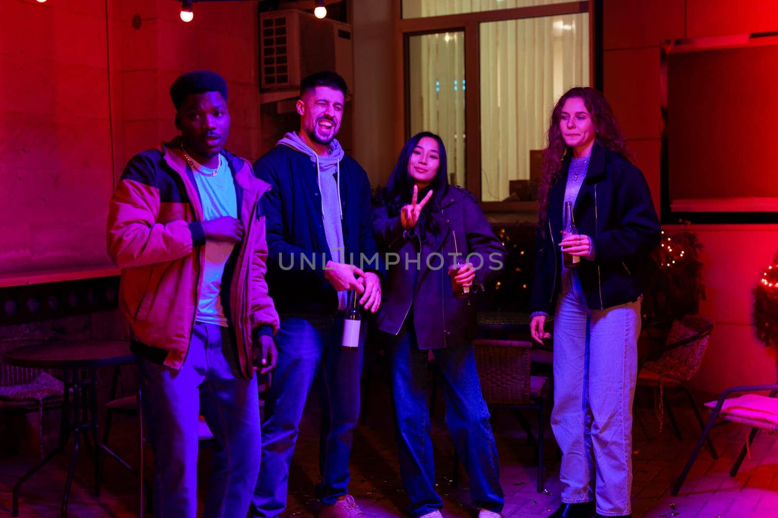 Friends gather for a fun night out in a warmly lit outdoor area. They hold drinks, share smiles, and enjoy each other's company under bright festive decorations.