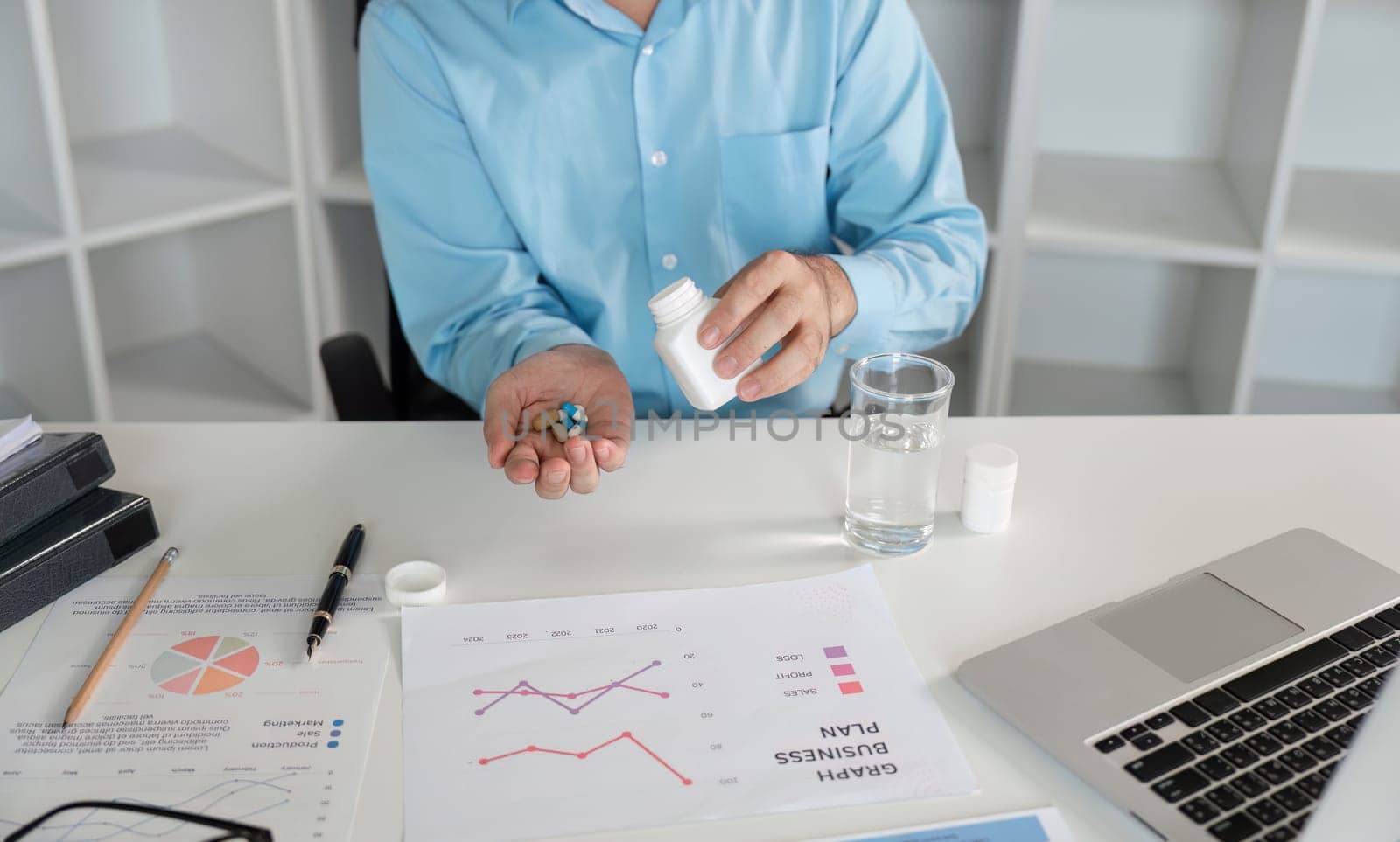 An office worker with a cold sits at a desk with medication, surrounded by business documents and a laptop, illustrating workplace illness.