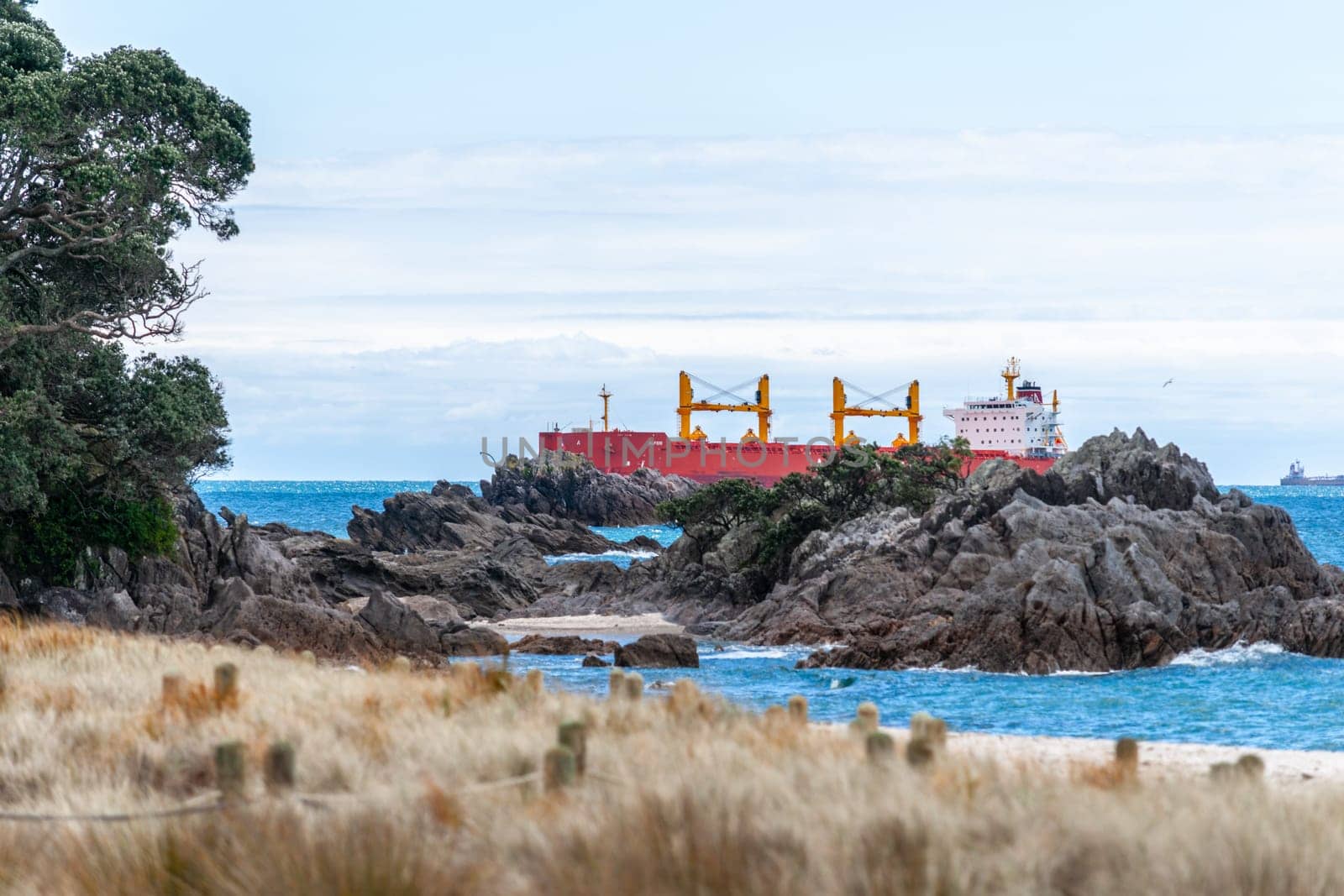 Mount Maunganui New Zealand - November 6 2012; Editorial-Red cargo ship passing rocky outcrop at base of Mount entering harbour port.