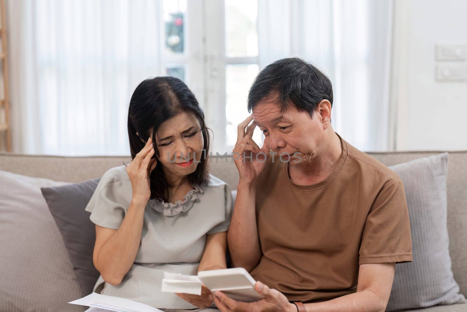 An elderly couple sitting on a couch, looking stressed while reviewing bills and financial documents at home.