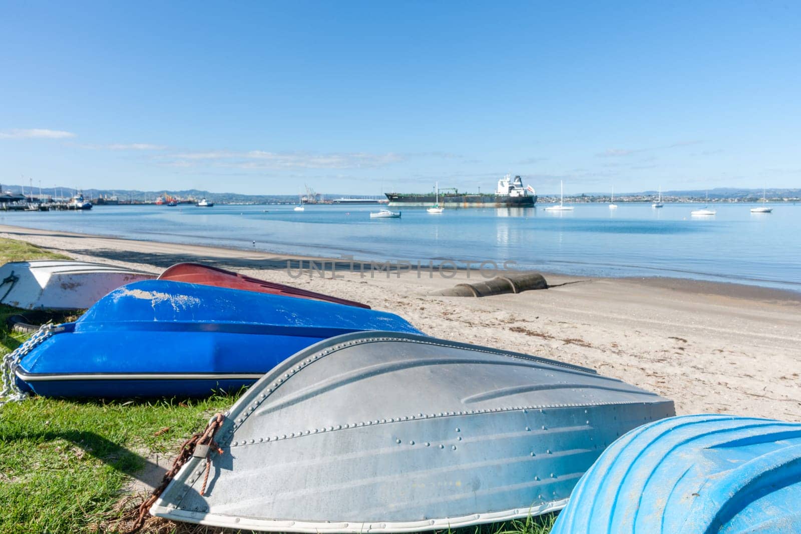Group of upturned dinghies pulled up on grass edge to beach with boats and port activity in background to Pilot Bay Mount Maunganui.