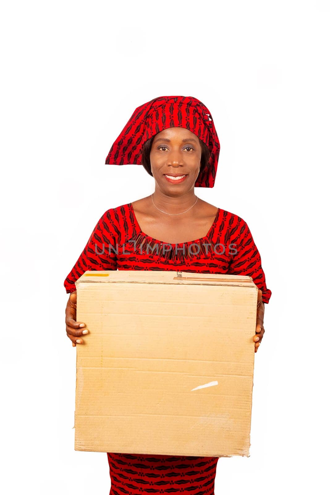 happy beautiful mature african woman standing and dressed in red loincloth suit holding a big cardboard box on white background