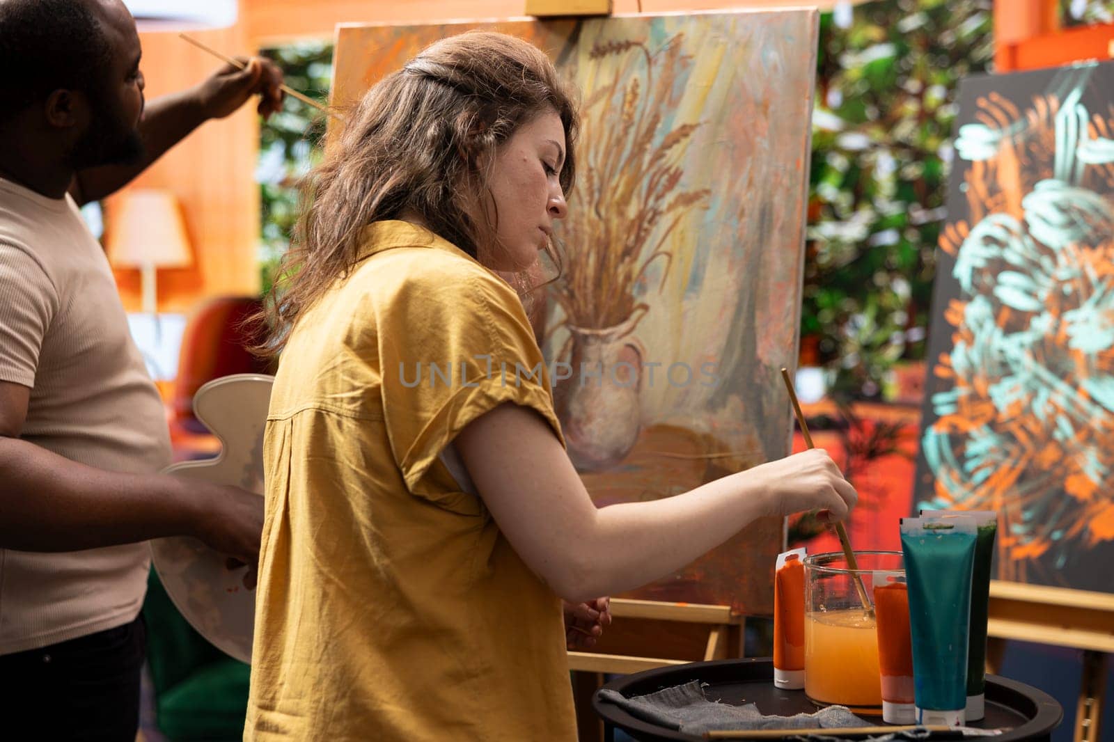 Team of artists painting a beautiful rustic creation on canvas, mixing colors together on a palette to create a nature inspired design. Diverse painters enjoy quality time in art studio.