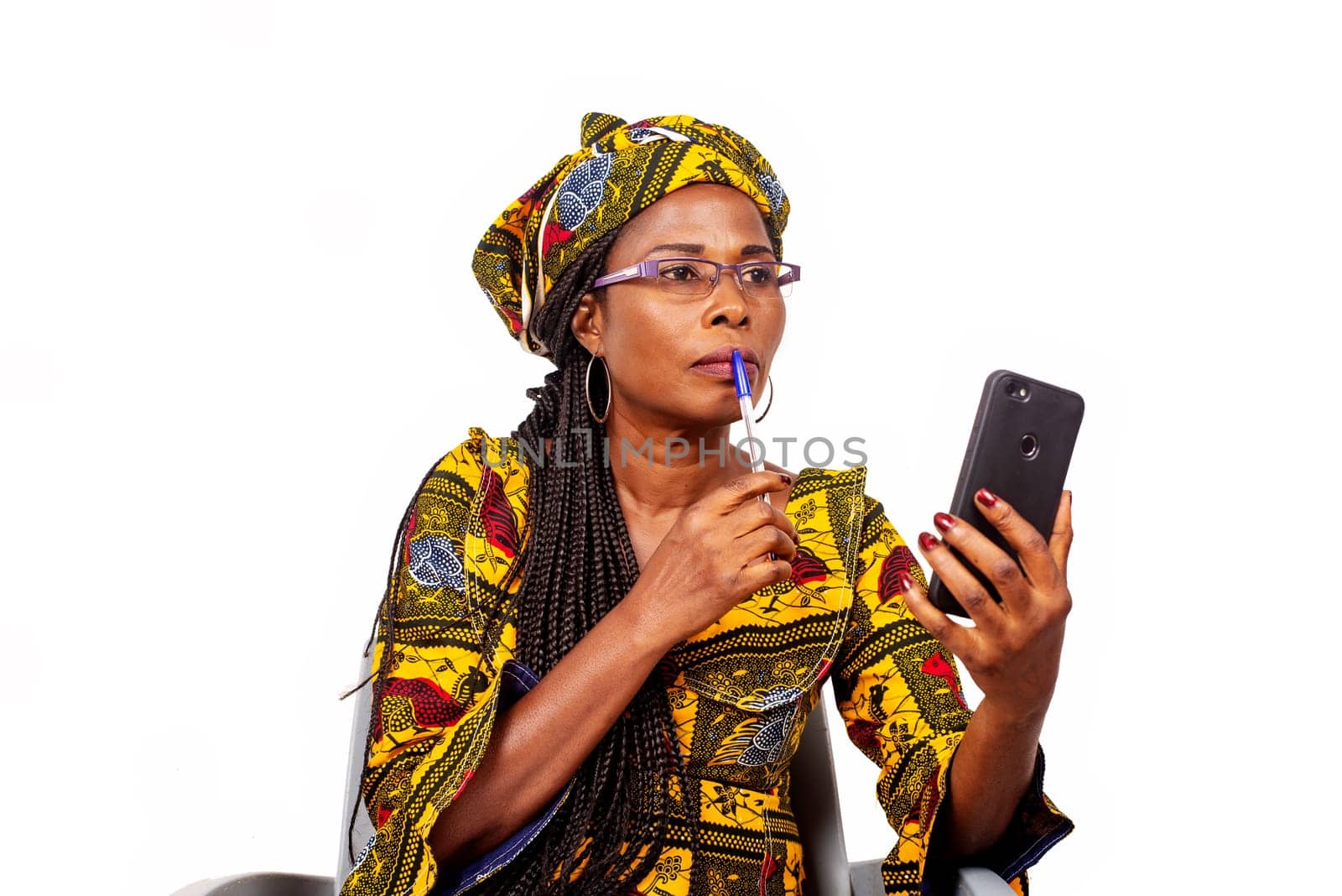 Beautiful adult woman in colorful loincloth is sitting holding a pen in her mouth, thinking while looking at the cell phone.