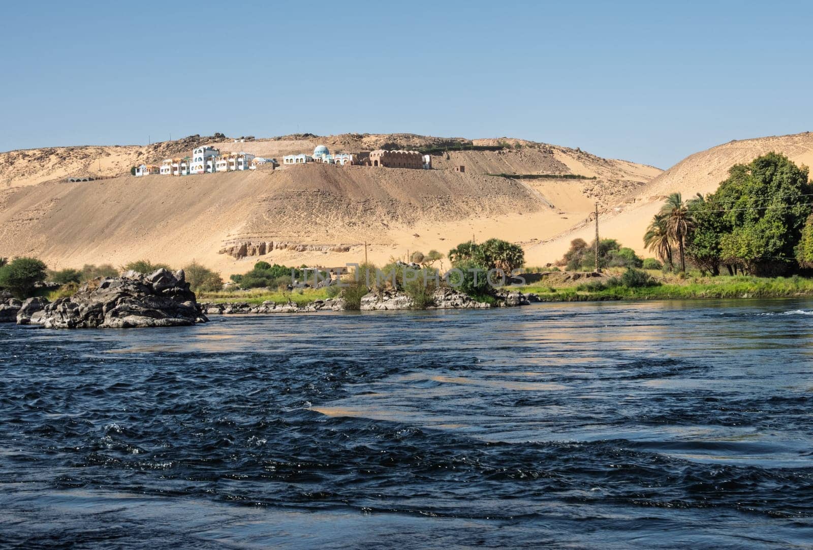 Granite rocks in the fast flowing water of the River Nile near Aswan Egypt with resort on hilltop