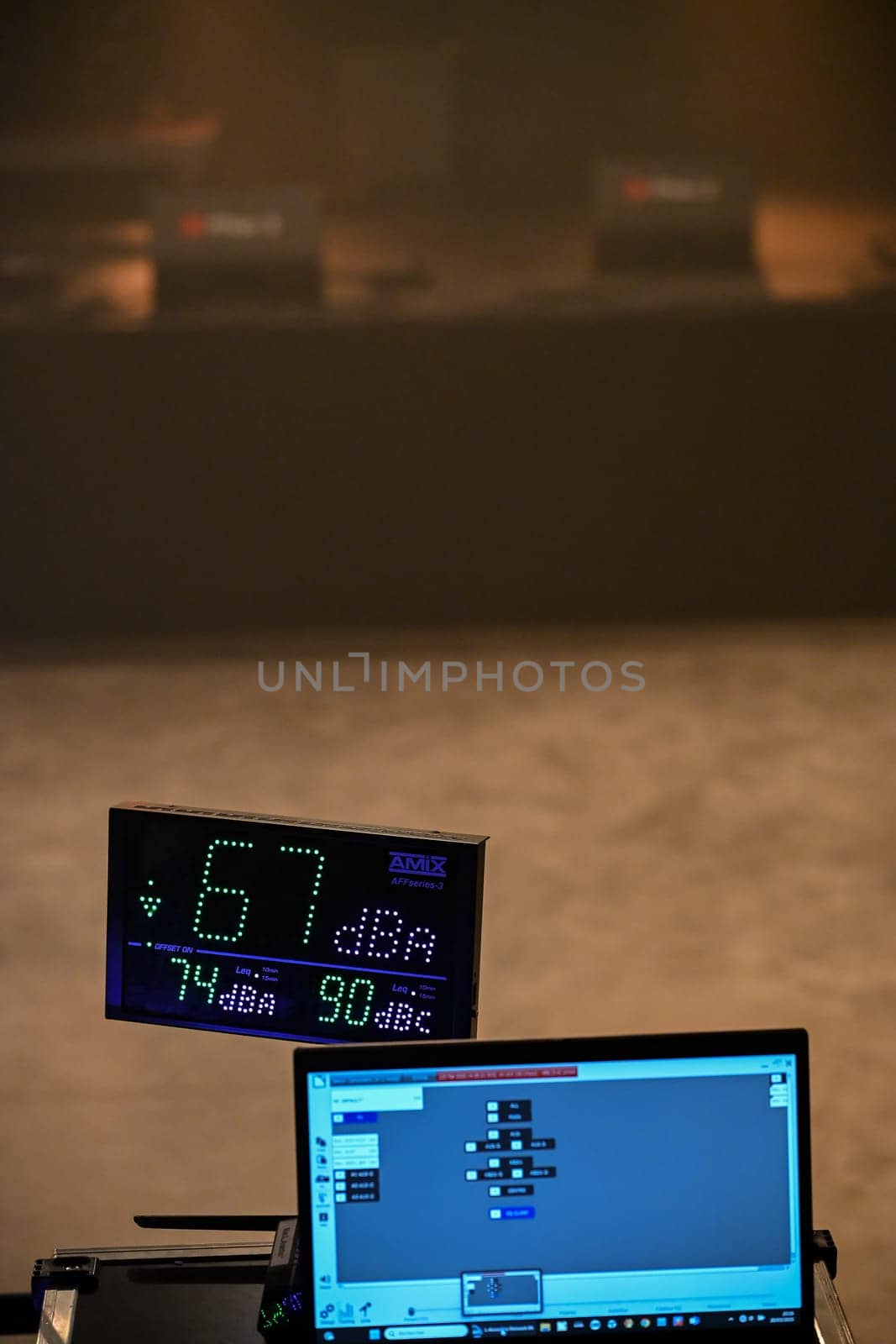 Sound level is monitored at a concert, showing 67 dBA on a digital display. A laptop runs software for tracking decibel levels as part of audio management.