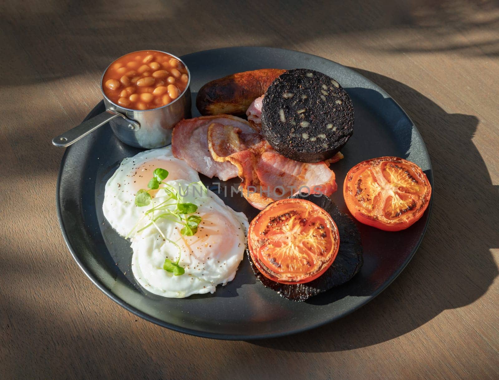 The full English Breakfast includes grilled bacon, sausage, baked beans, mushroom, tomatoes, black pudding is a sausage made with pork and flavored with pig blood and two hen's fried eggs with a runny yolk and watercress served on ceramic plate. use it as your Wallpaper, Poster and Space for text, Selective focus.