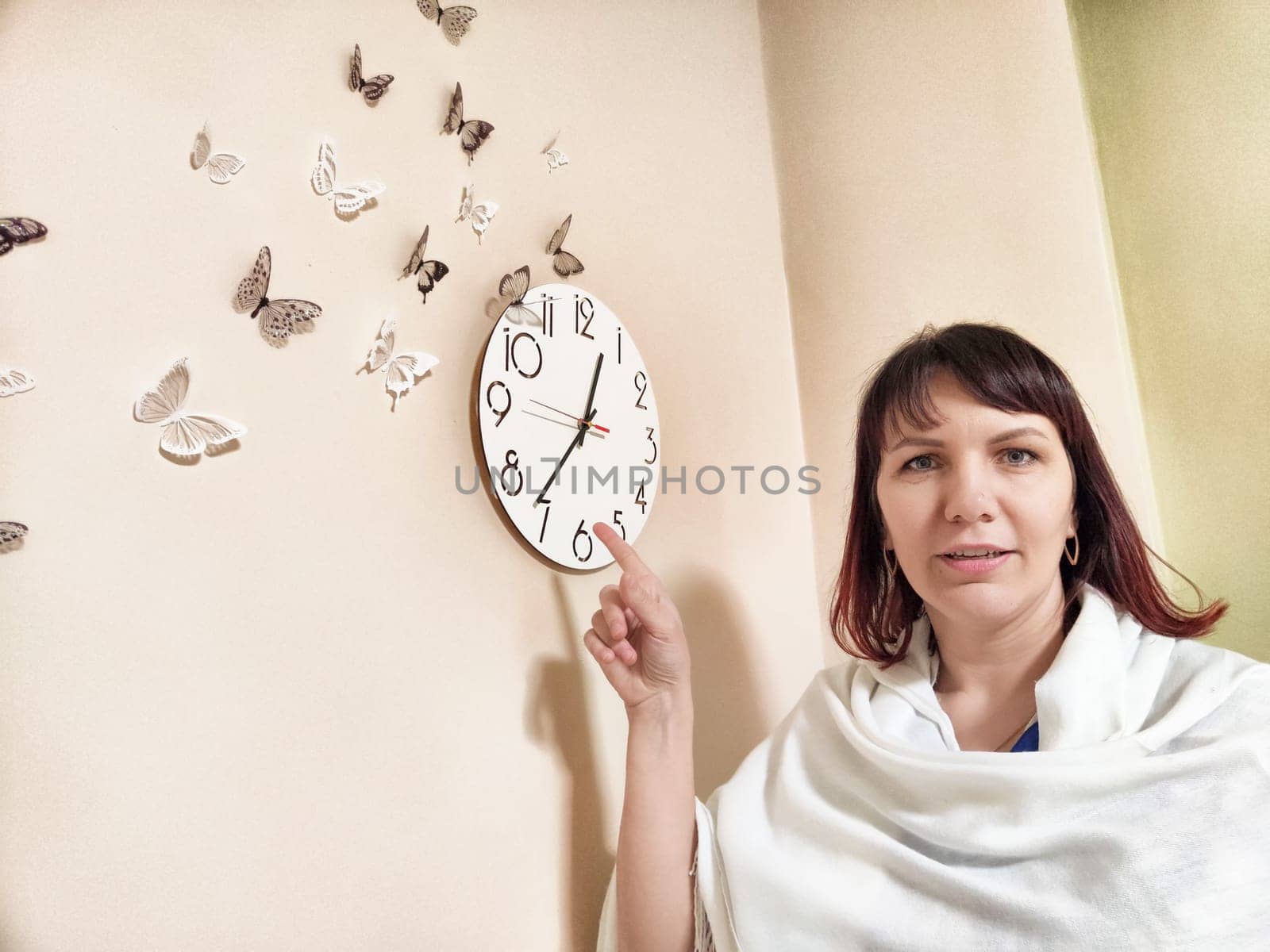 A woman psychologist in a cozy office points at a clock on the wall decorated with butterflies, indicating a time for patients.