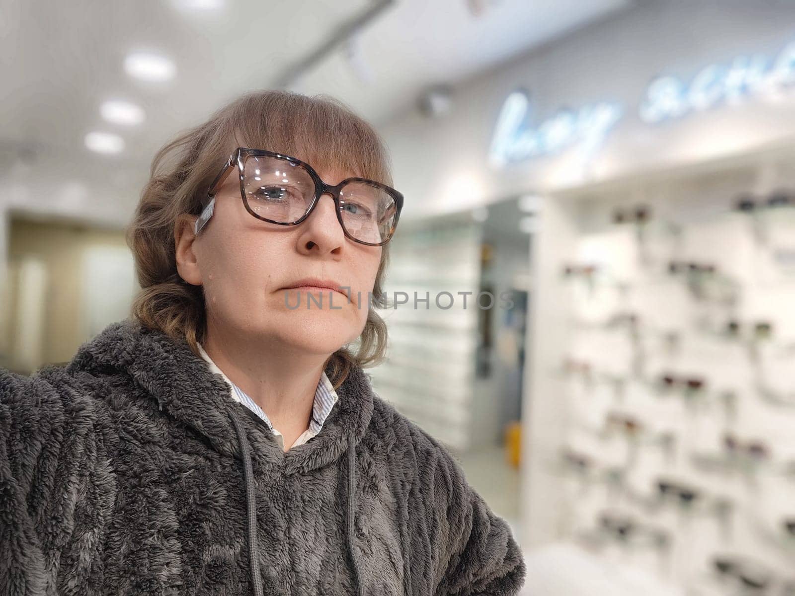 Woman with blonde hair trying on bold glasses in an optics store, capturing her experience in a selfie while browsing options.