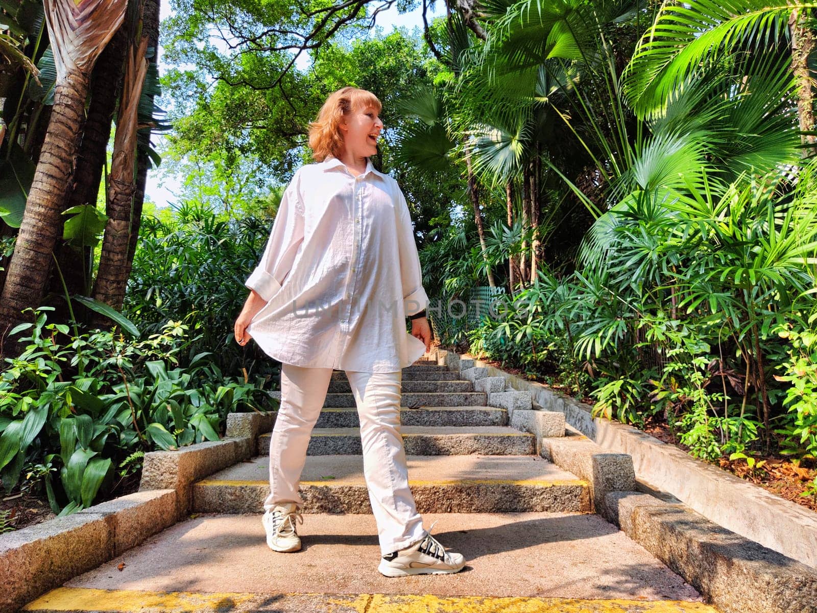 Mature woman pauses on a stone staircase in a vibrant, green setting, soaking in the sunshine while taking a casual selfie.