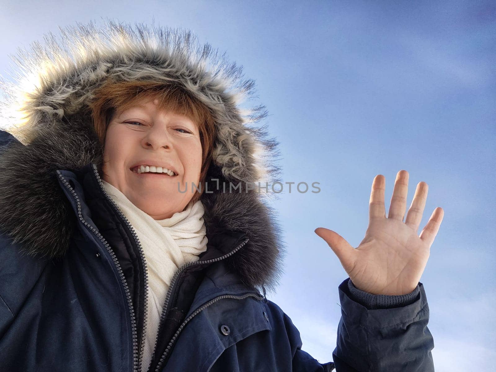 A middle aged woman in winter attire smiles and waves while capturing a joyful moment in a snowy location.