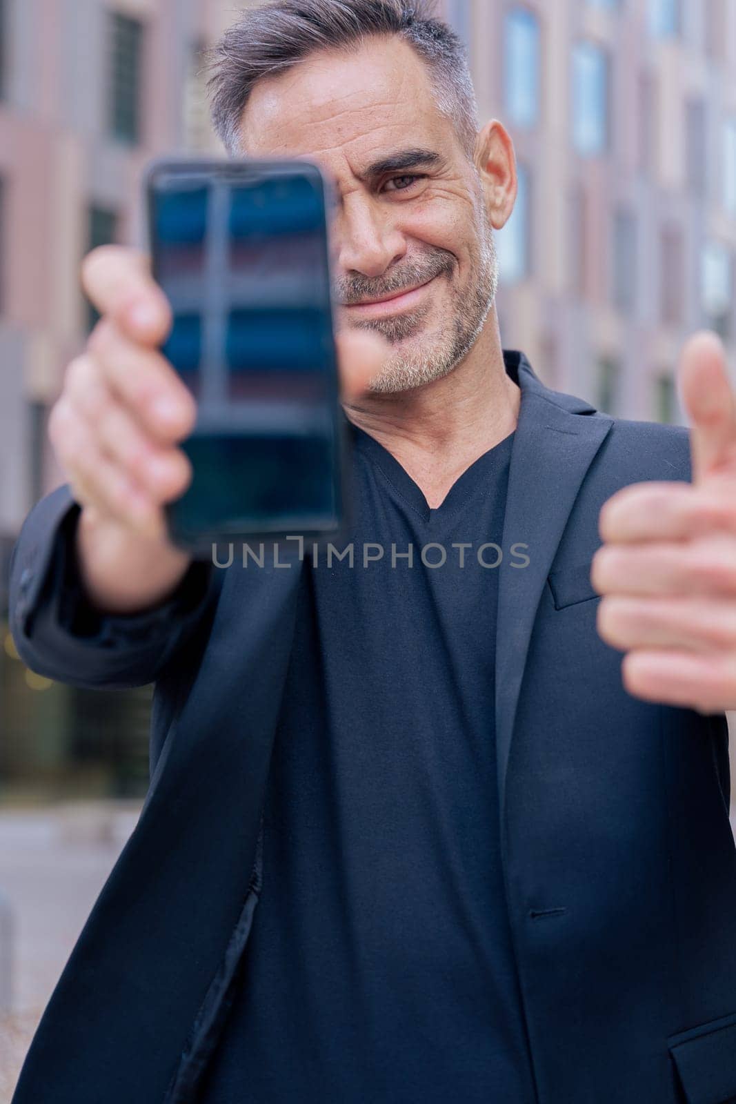 Smiling man holding smartphone towards the camera and giving a thumbs up outdoors. He wears a black jacket and stands in a modern urban space. Concept of approval, technology, and confidence. Vertical