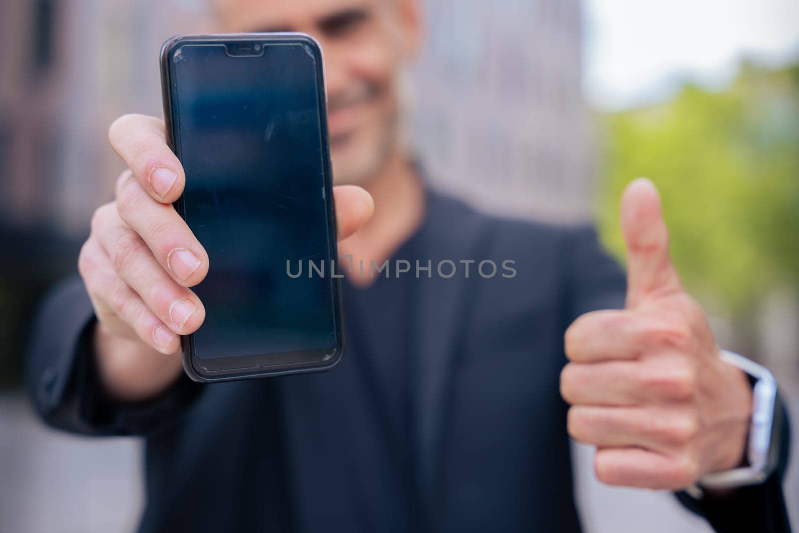 Smiling man holding smartphone towards the camera and giving a thumbs up outdoors. He wears a black jacket and stands in a modern urban space. Concept of approval, technology, and confidence.