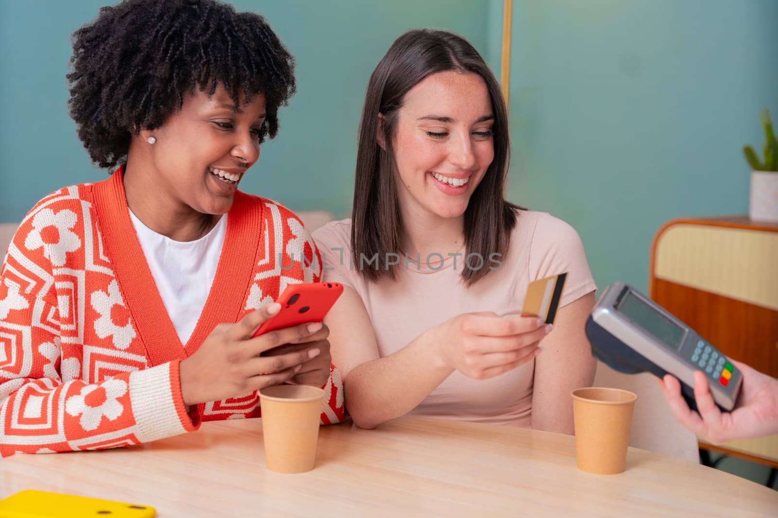 African American and Caucasian women smiling while paying at a coffee shop. One holds a card, the other a phone. Concept of friendship, technology, and casual lifestyle in a modern setting.