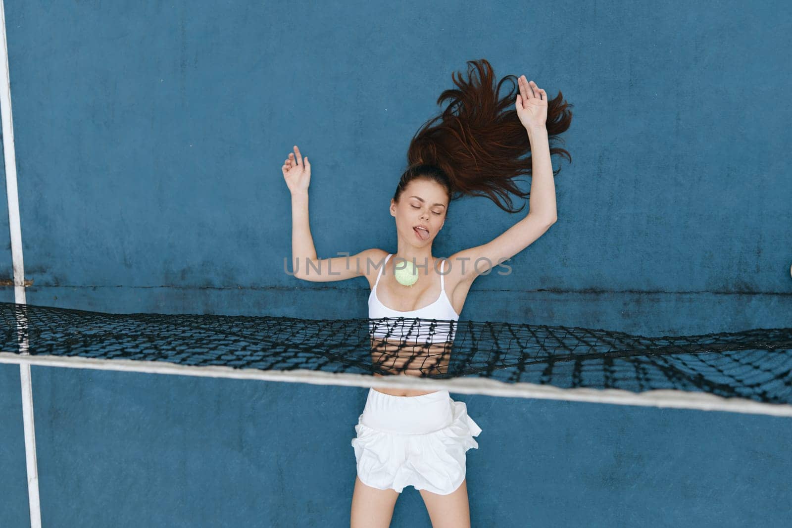 Energetic young woman in a trendy white ensemble, joyfully interacting with a tennis ball, radiating summer vibes and enthusiasm against a striking blue backdrop