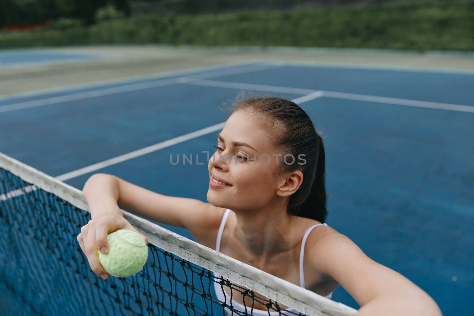 A young female athlete leans on the tennis net, holding a bright yellow tennis ball Her expression radiates happiness and determination, set against a vibrant blue tennis court, ideal for promoting an active lifestyle