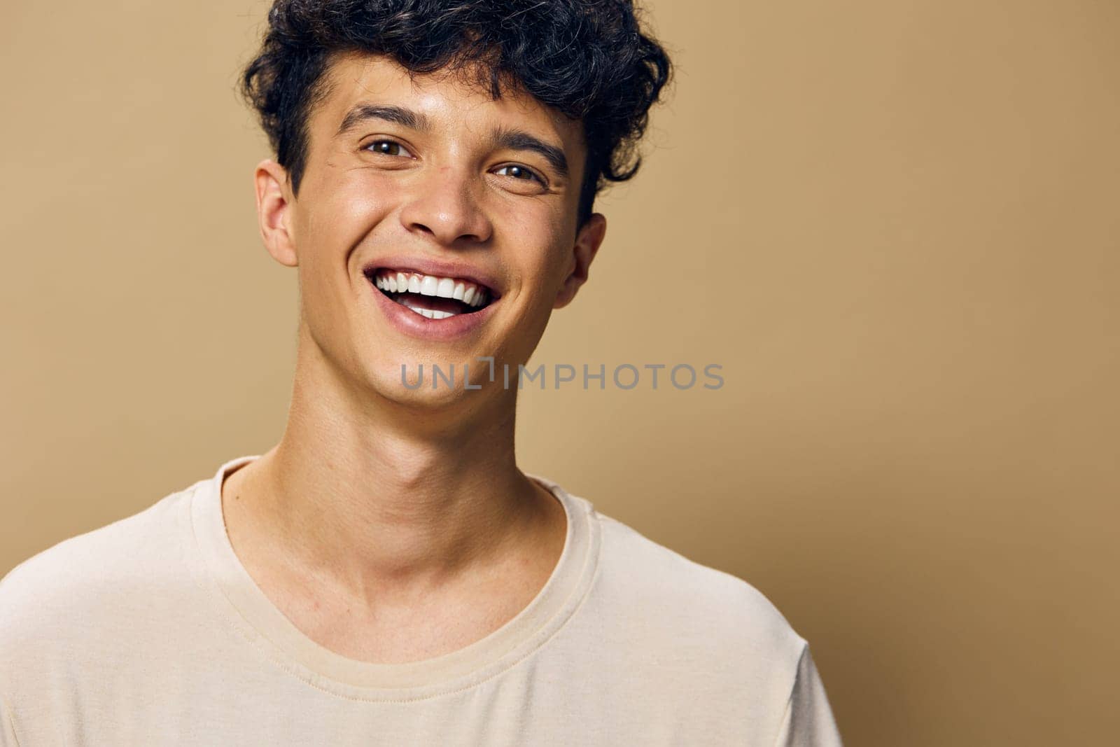A cheerful young male with curly hair, joyfully smiling against a warm beige background, portraying happiness and friendliness, perfect for lifestyle, emotions, and youth concepts