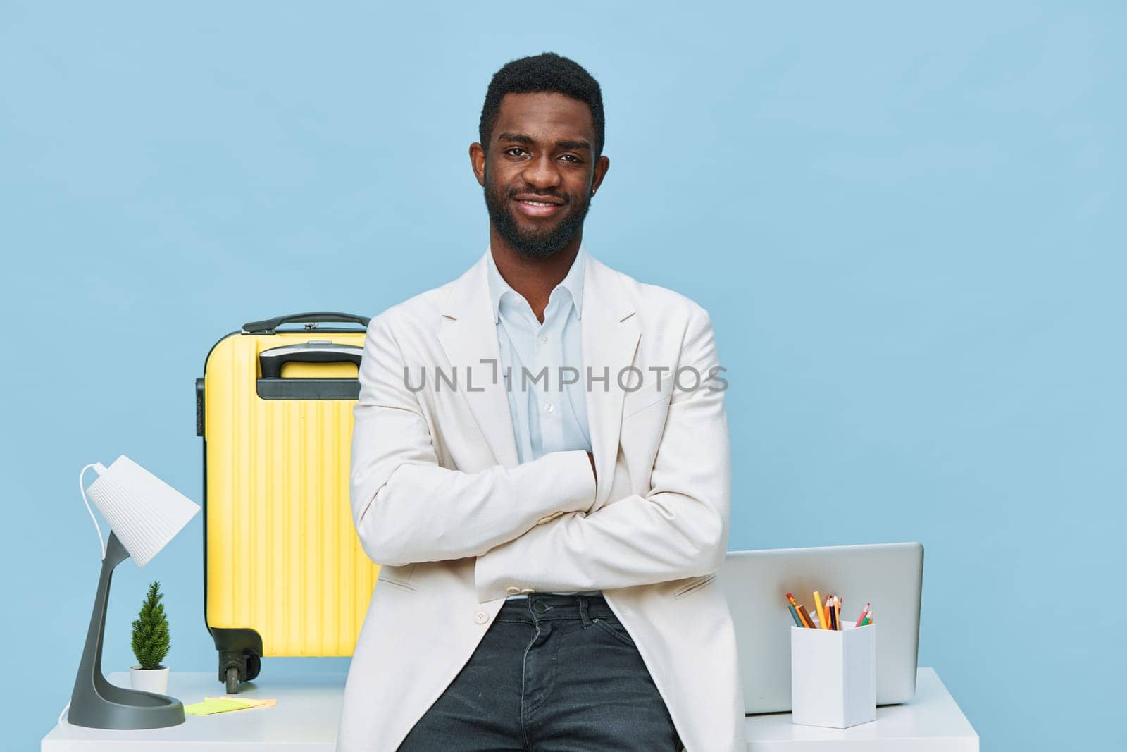 Happy professional man with dark skin, curly hair, and casual light blazer, standing confidently with arms crossed in modern workspace with laptop, bright yellow suitcase, desk lamp, and office accessories on light blue background, studio shot. Business lifestyle concept