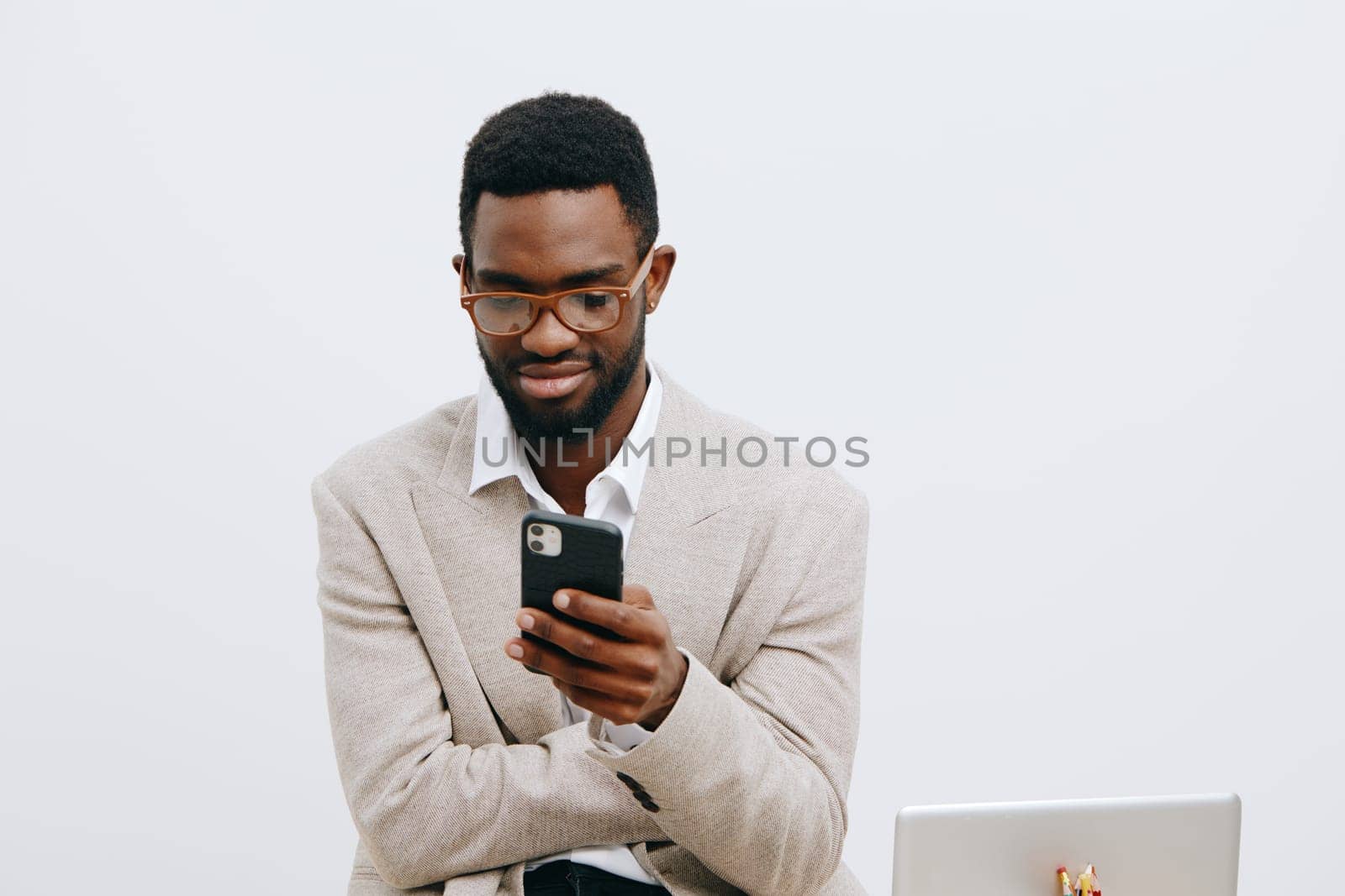 Professional young man with glasses engaging with his mobile device at work, wearing a casual blazer, standing against a plain white background, demonstrating tech-savvy, communication skills, modern workspace, digital age, flexible working concept.
