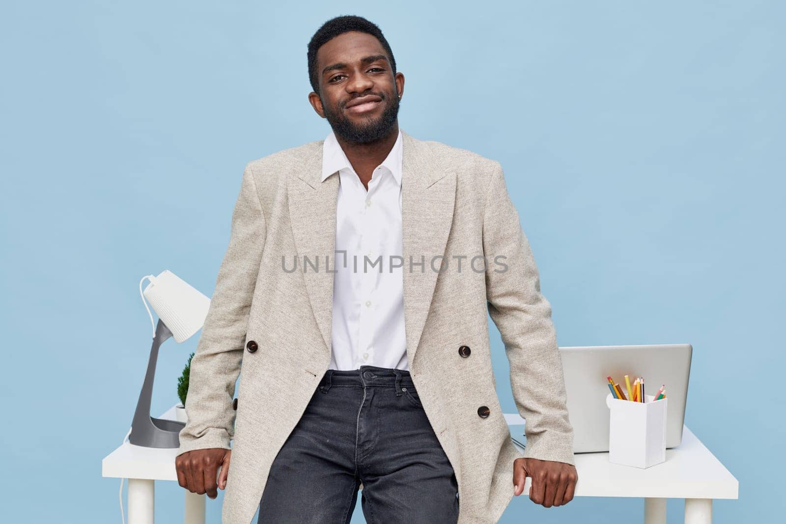 Cheerful man with dark skin and curly hair, dressed in a beige blazer and white shirt, posing confidently in a stylish office with computer, stationery, and bright background, conveying success and positivity, workplace concept.