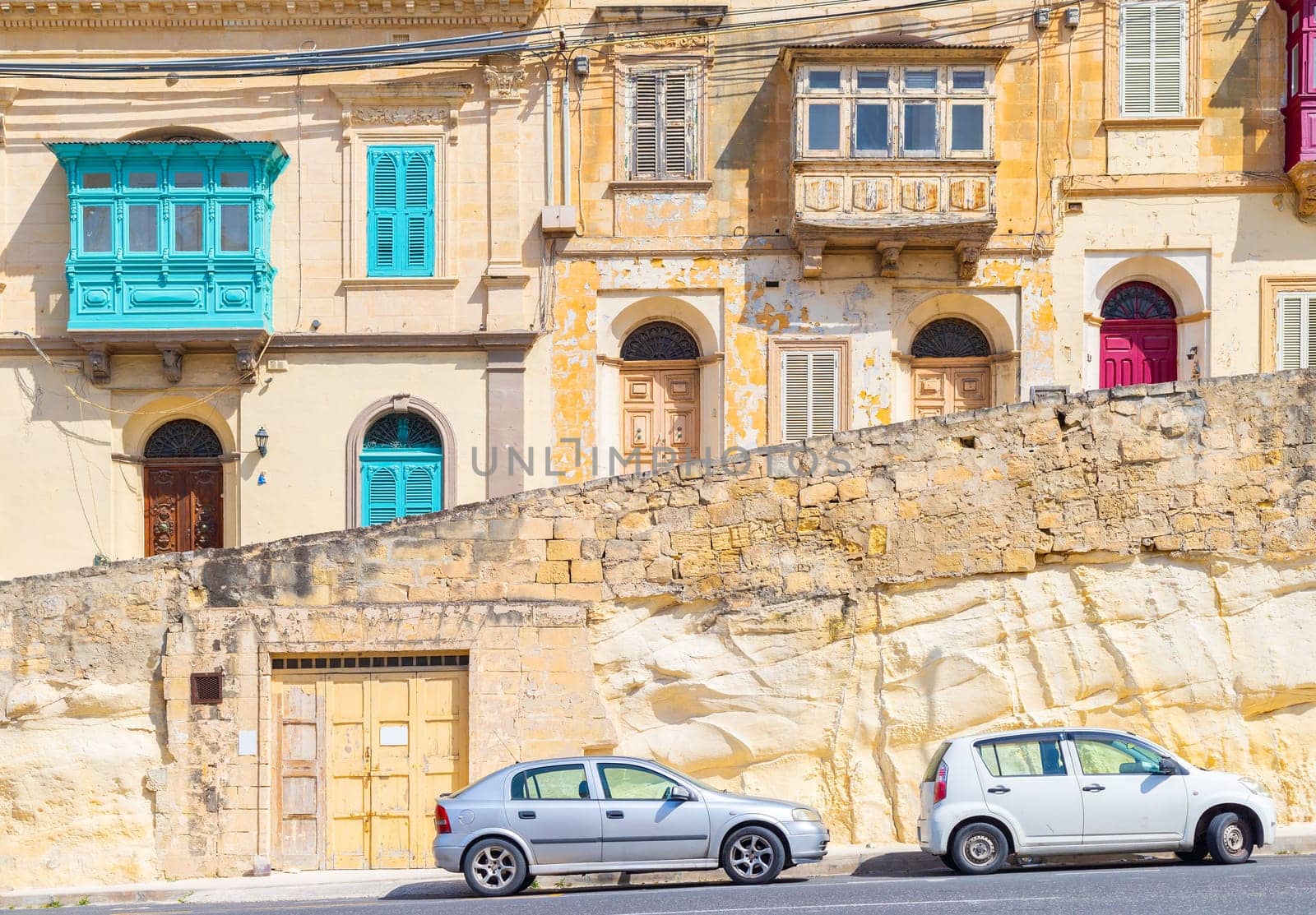 View of a typical maltese street with houses in traditional architecture style with colorful doors, balconies, windows and parked cars.