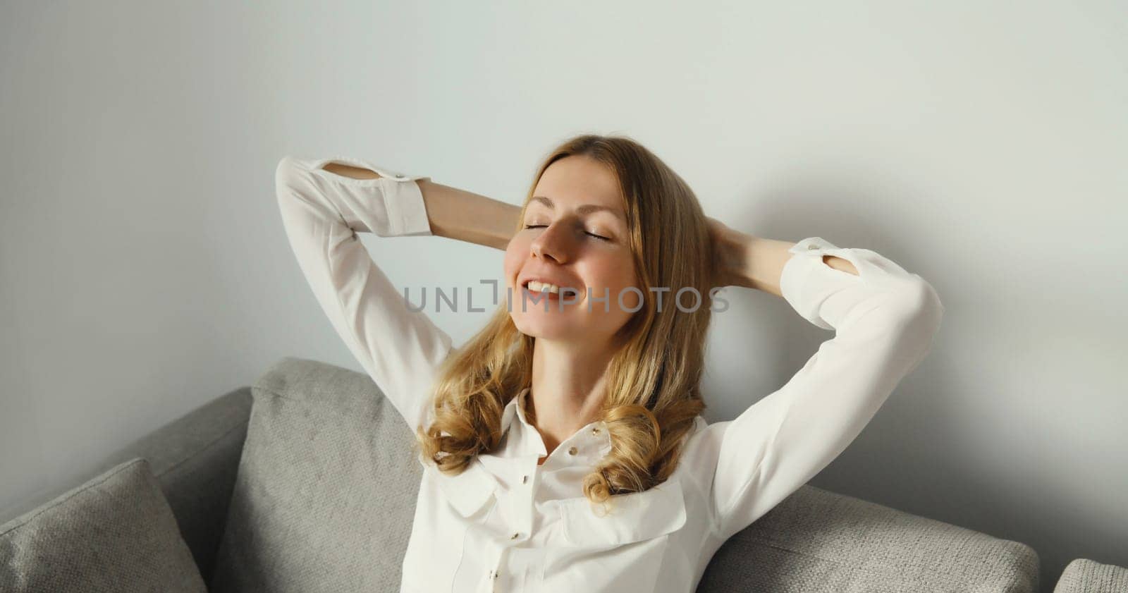 Happy modern relaxed young woman enjoying at home, satisfied girl resting with closed eyes and folded hands behind the head sitting on the couch in living room